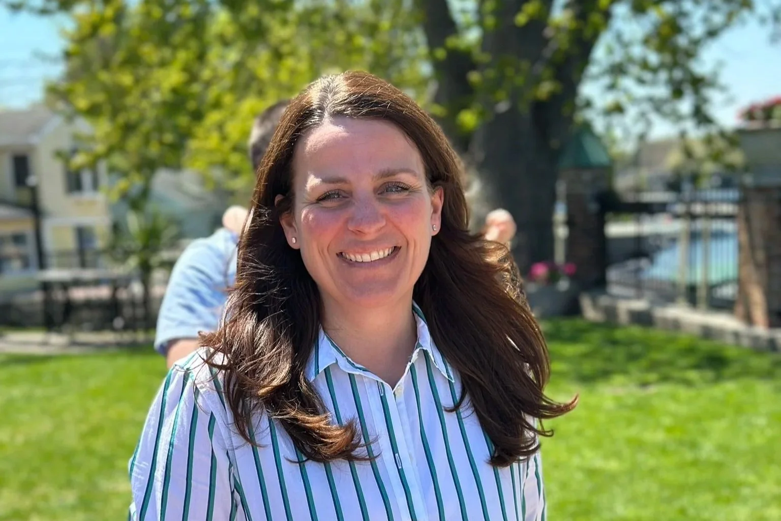 A woman smiling outdoors, wearing a white and green striped shirt, with a blurred background of trees, grass, and a building.