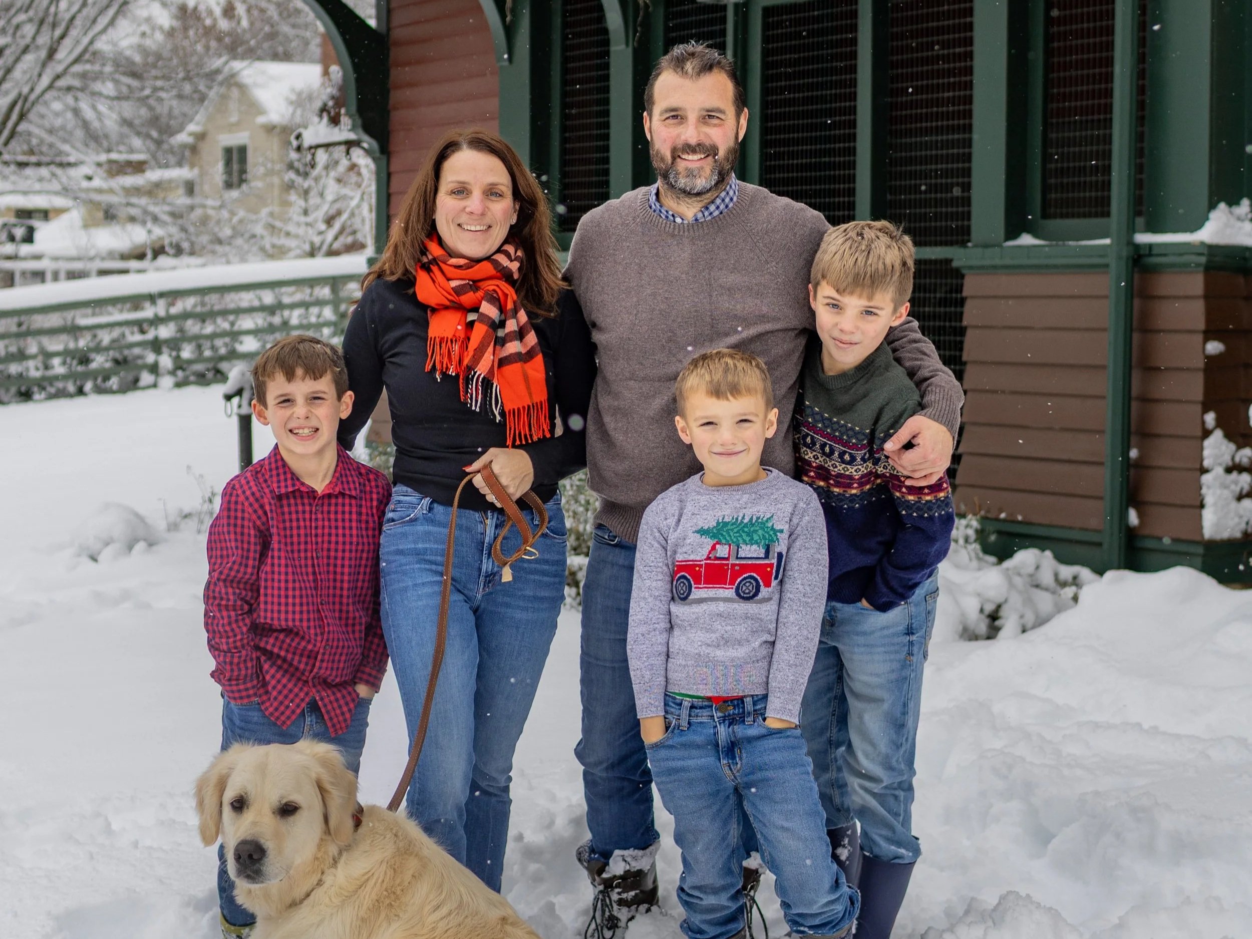 A family of six, including two adults and four boys, along with a golden retriever, standing outdoors in snowy weather in front of a house. The family is dressed in winter clothing and smiling.
