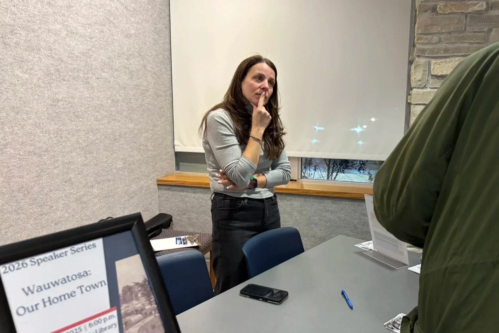 A woman with long brown hair, wearing a gray shirt and black pants, standing in a room with beige walls, a window with a white shade, and a brick accent wall. She has her right hand resting on her chin, appearing thoughtful or listening. Partially vi