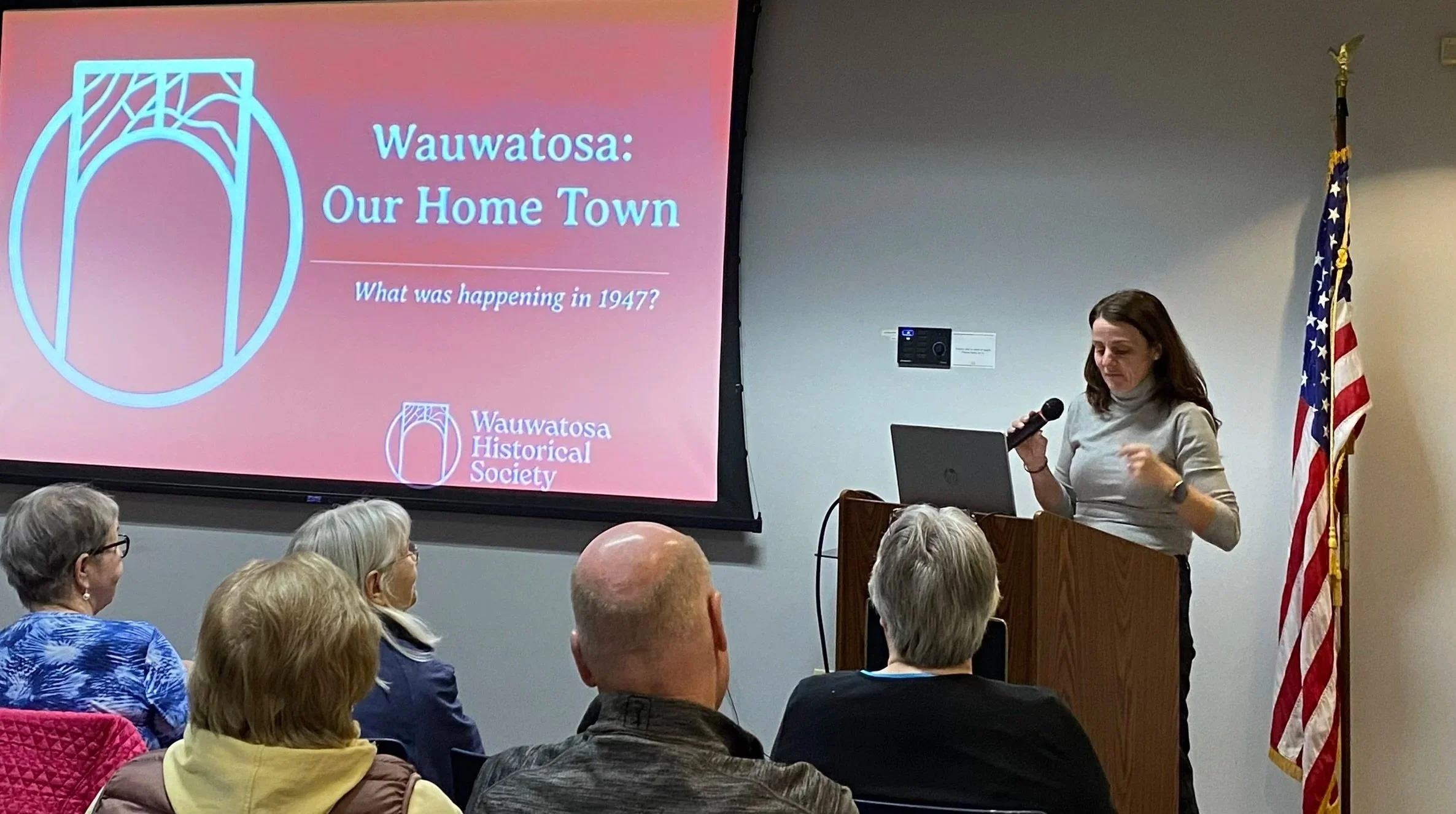 A woman giving a presentation at the Wauwatosa Historical Society, with a projection screen displaying the title 'Wauwatosa: Our Home Town' and the question 'What was happening in 1947?', and an American flag standing beside her.
