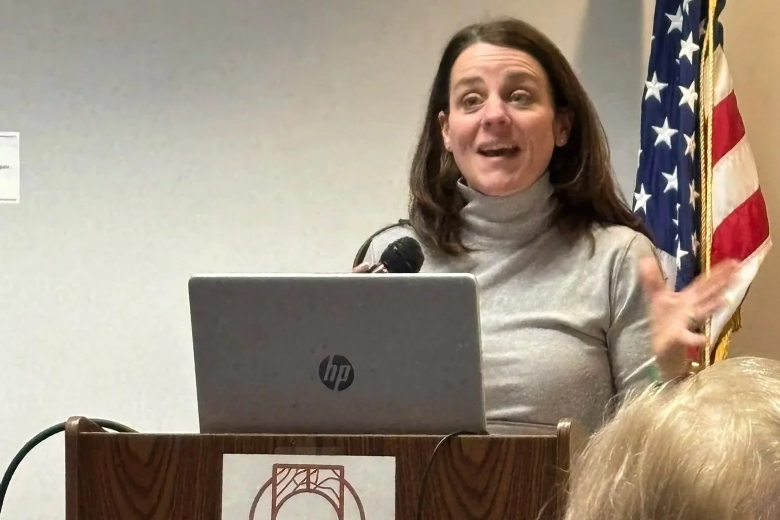 A woman with shoulder-length brown hair speaking at a podium with a laptop, American flag in the background, gesturing with her hand, wearing a light gray turtleneck.