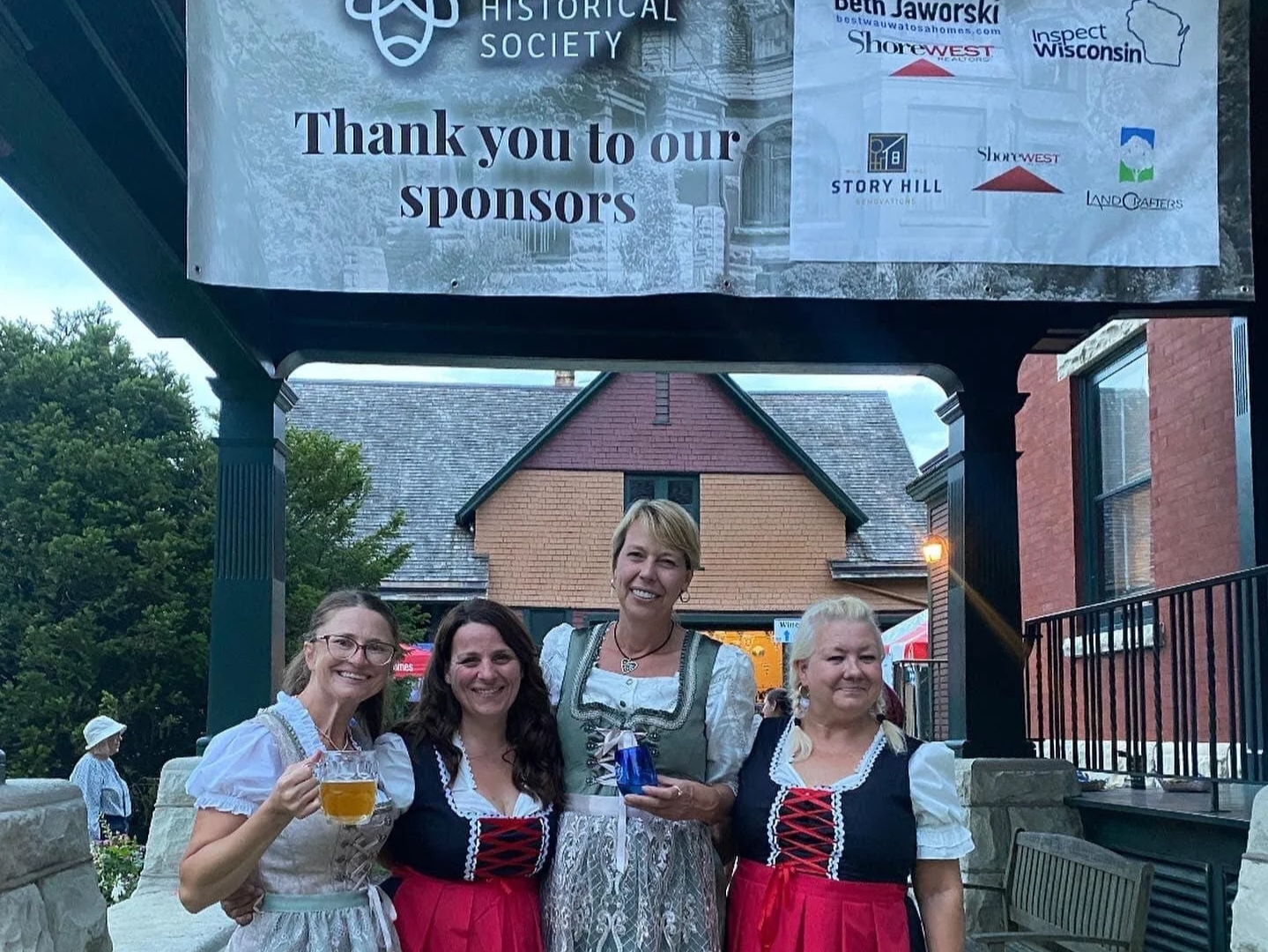 Four women in traditional dresses standing outdoors, smiling, with a building and trees in the background. One woman is holding a mug of beer.