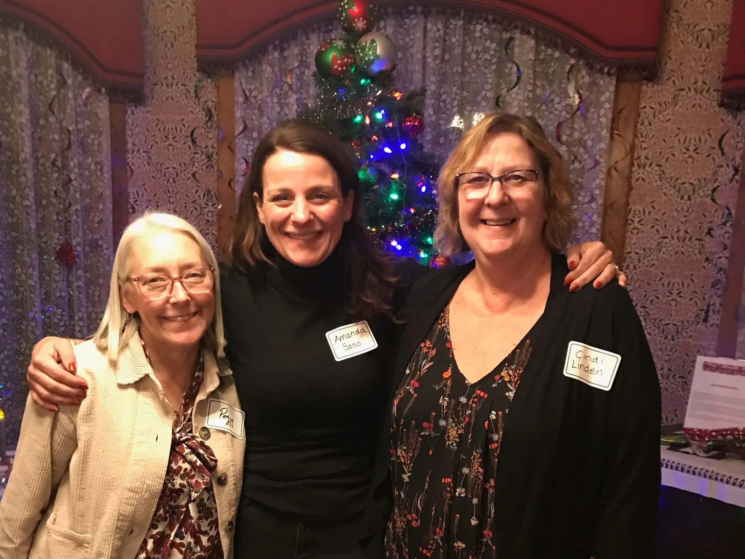 Three women smiling and standing together in front of a Christmas tree, with lights and ornaments, at a holiday gathering. They are wearing name tags.