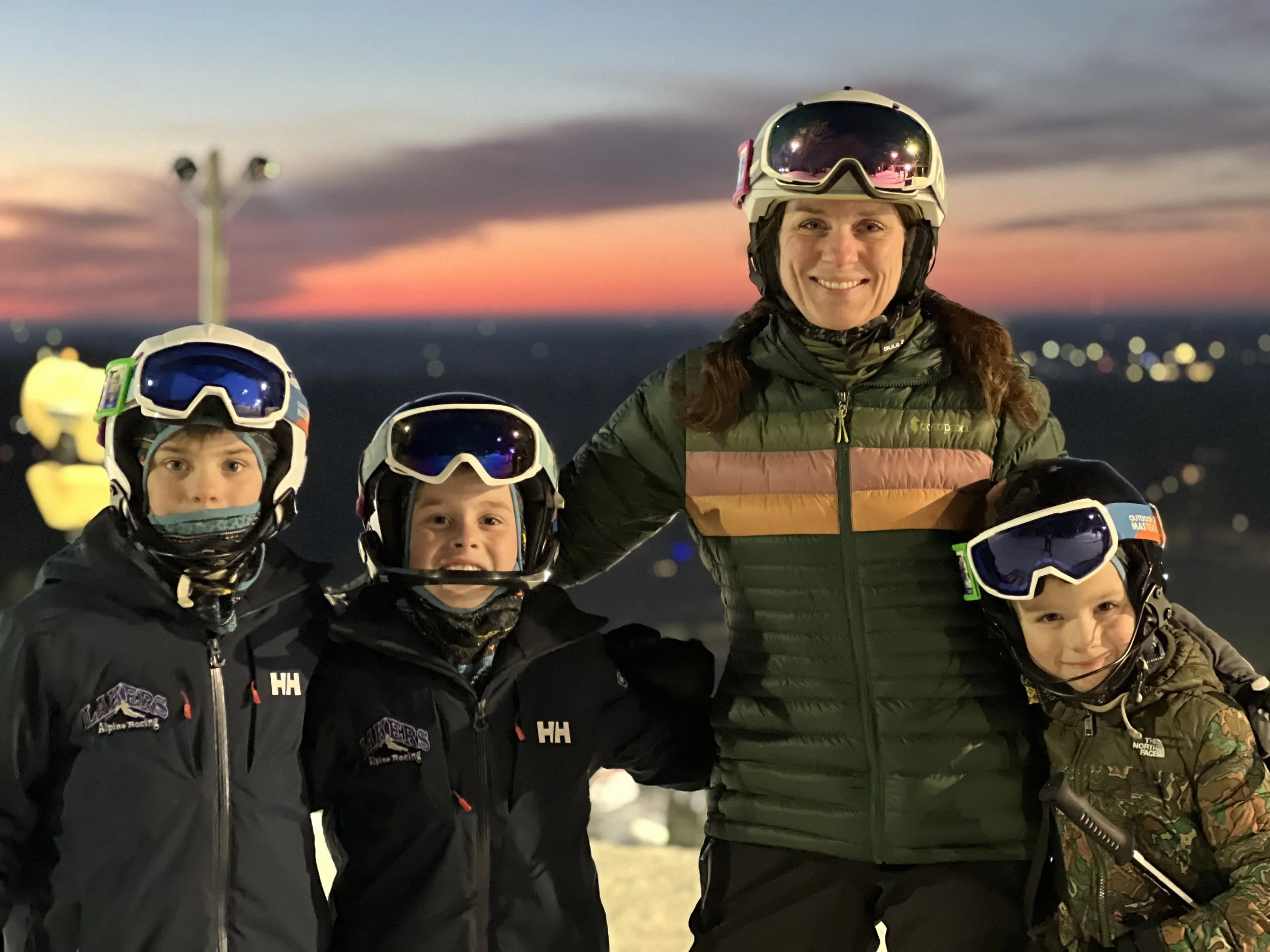 A group of four children and an adult woman in winter sports gear, wearing helmets and goggles, standing outdoors at dusk with a sunset sky in the background.