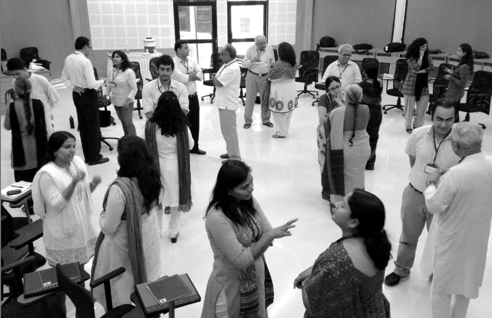 A black and white photo of about 20 people talking to one another in pairs. They are in an open, office-style space with chairs scattered on the periphery
