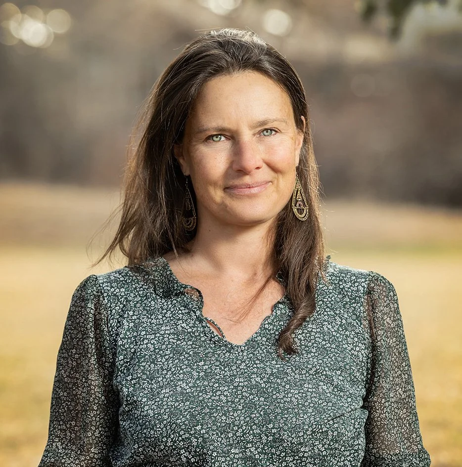 A woman with long brown hair and blue eyes wearing a green patterned blouse and gold earrings outdoors on a sunny day.