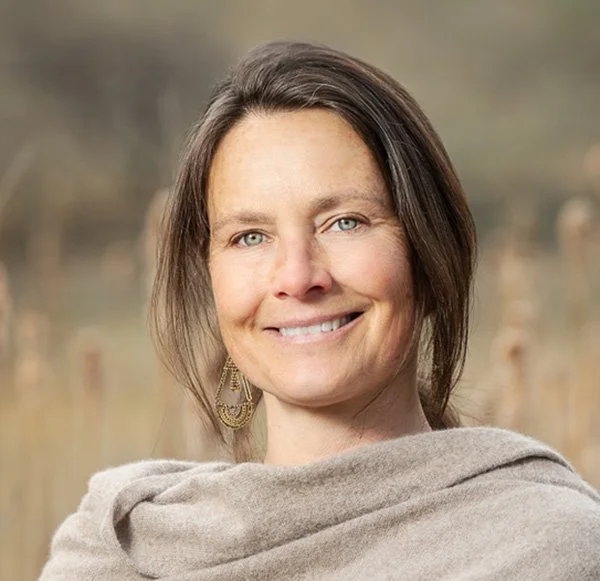 A woman with shoulder-length brown hair and blue eyes, smiling outdoors with a blurred natural background, wearing a beige shawl and gold earrings.