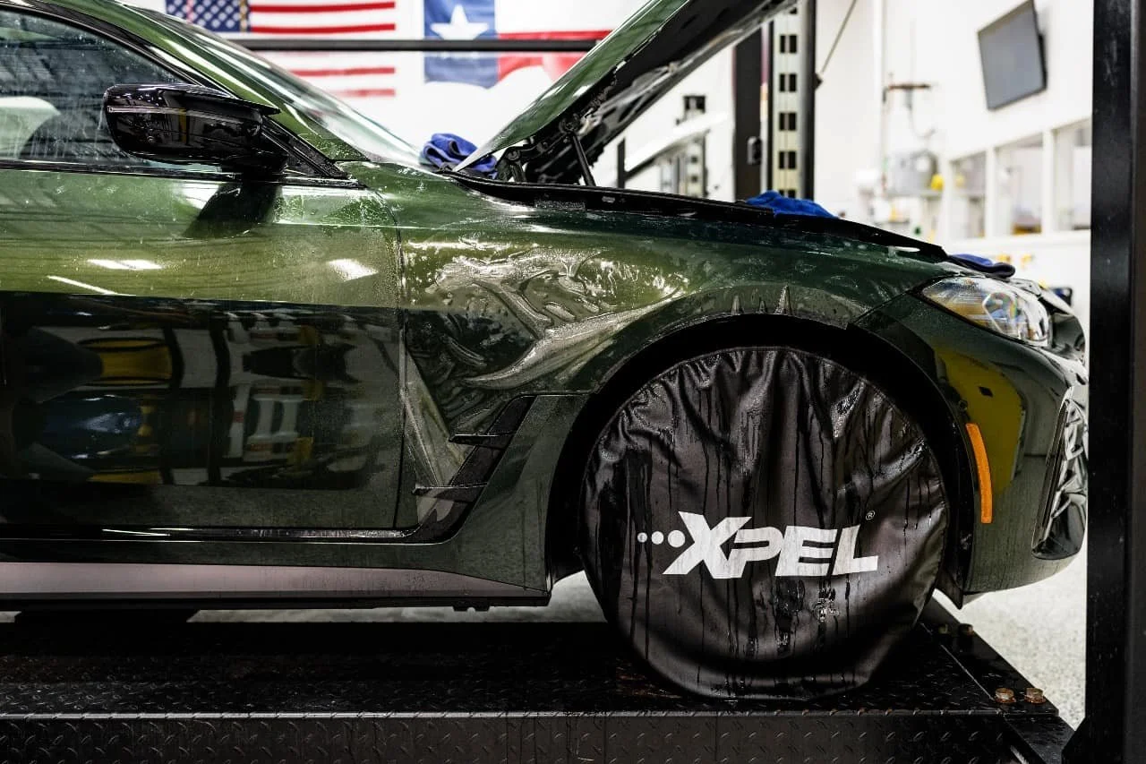 Close-up of a green car with front fender damage and a tire covered with a black protective cover labeled 'XPEL' inside a garage or workshop.