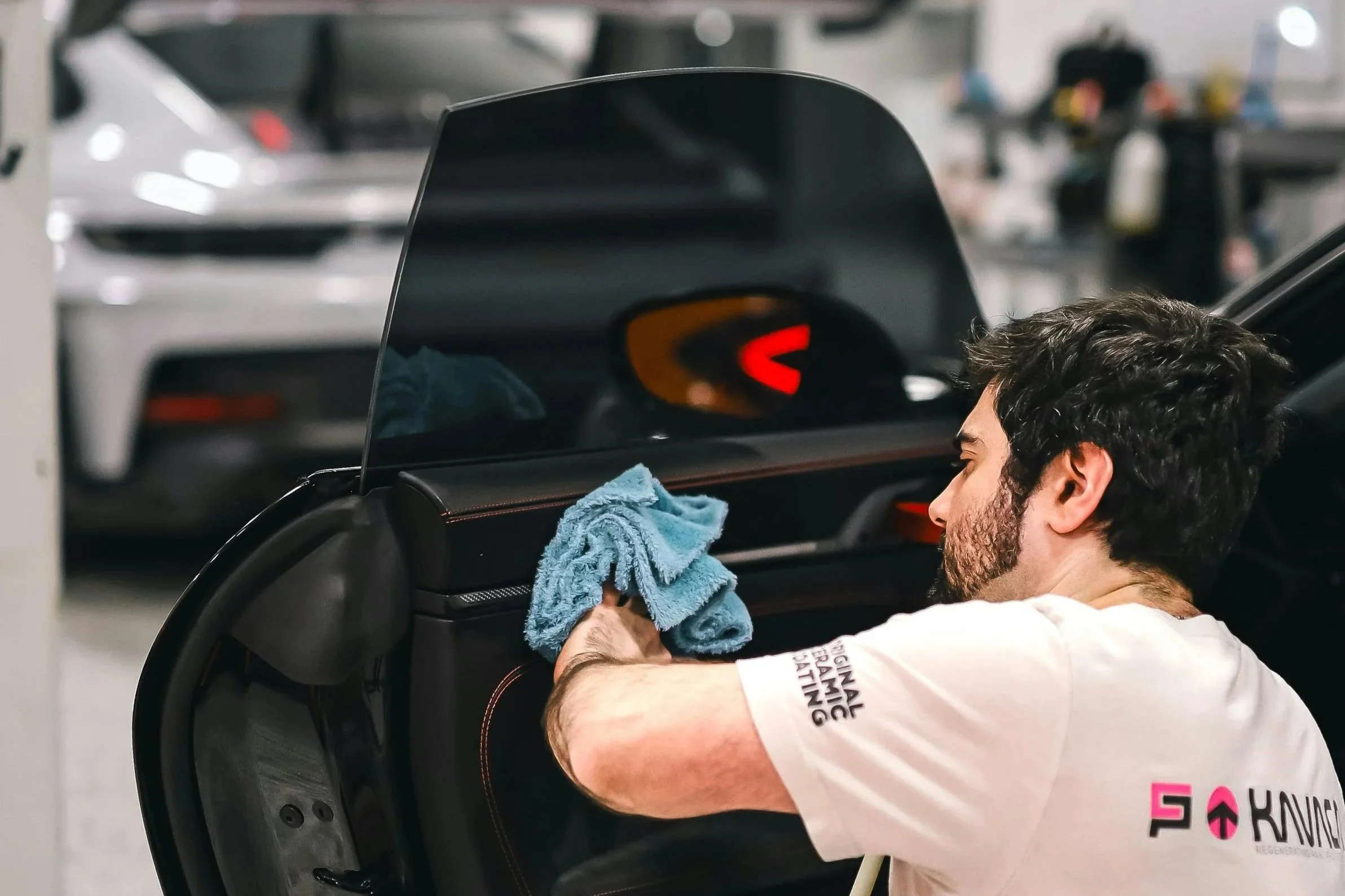 A man cleaning the interior of a car with a blue cloth in a garage. Multiple vehicles are visible in the background.