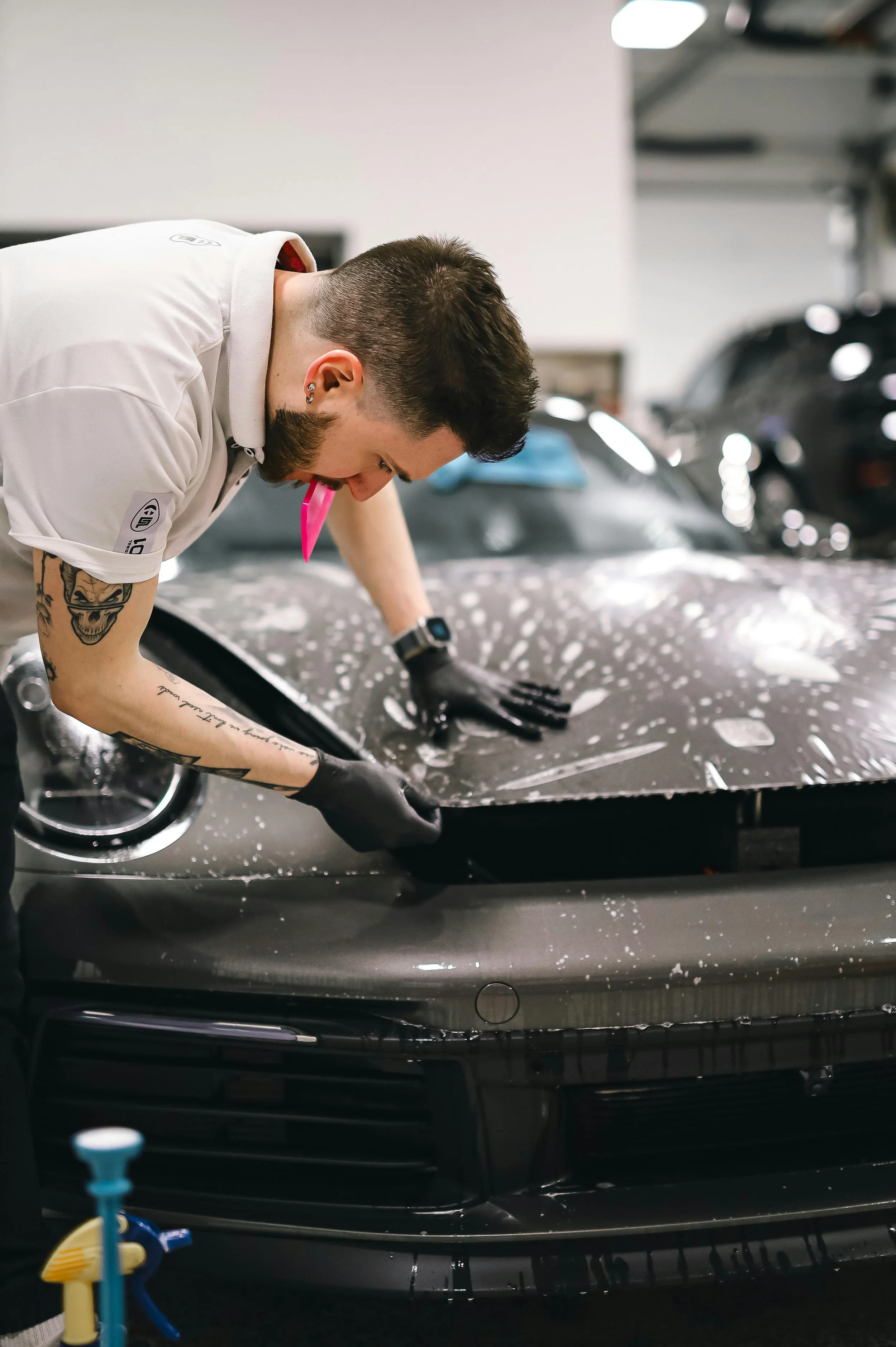 A man wearing black gloves is applying a self-adhesive wrap to the hood of a black luxury car, in an indoor garage. The man has short dark hair, tattoos on his arms, and is wearing a white polo shirt.