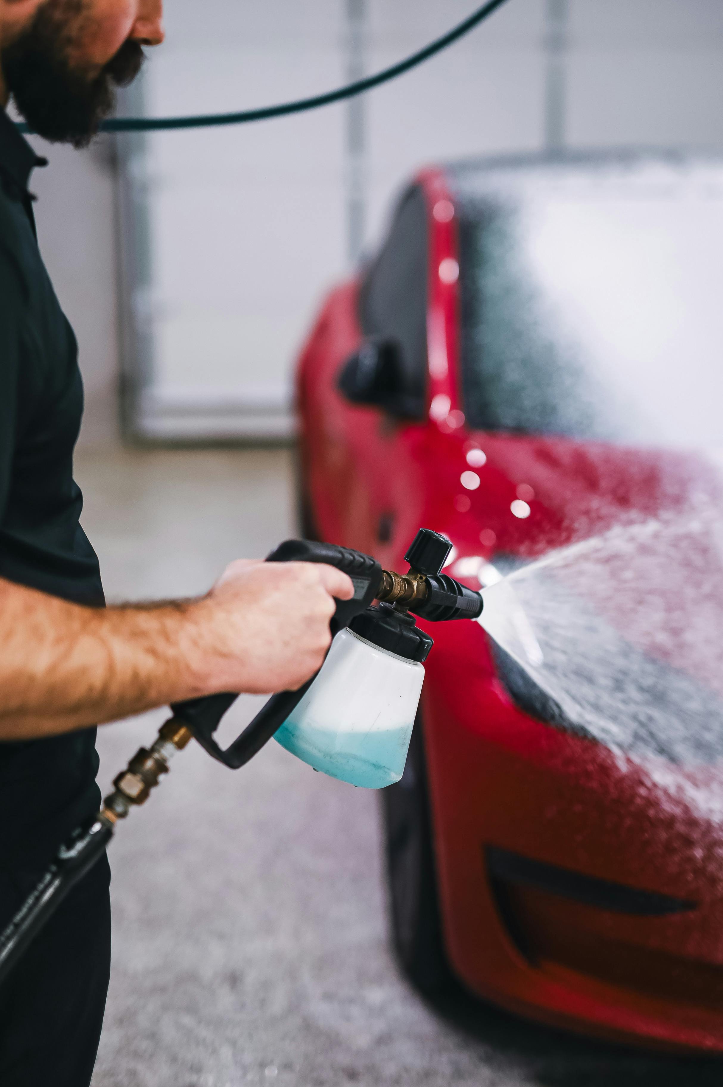 Person using a pressure washer to clean a red car, focusing on the person's arm and the nozzle spraying water on the vehicle.
