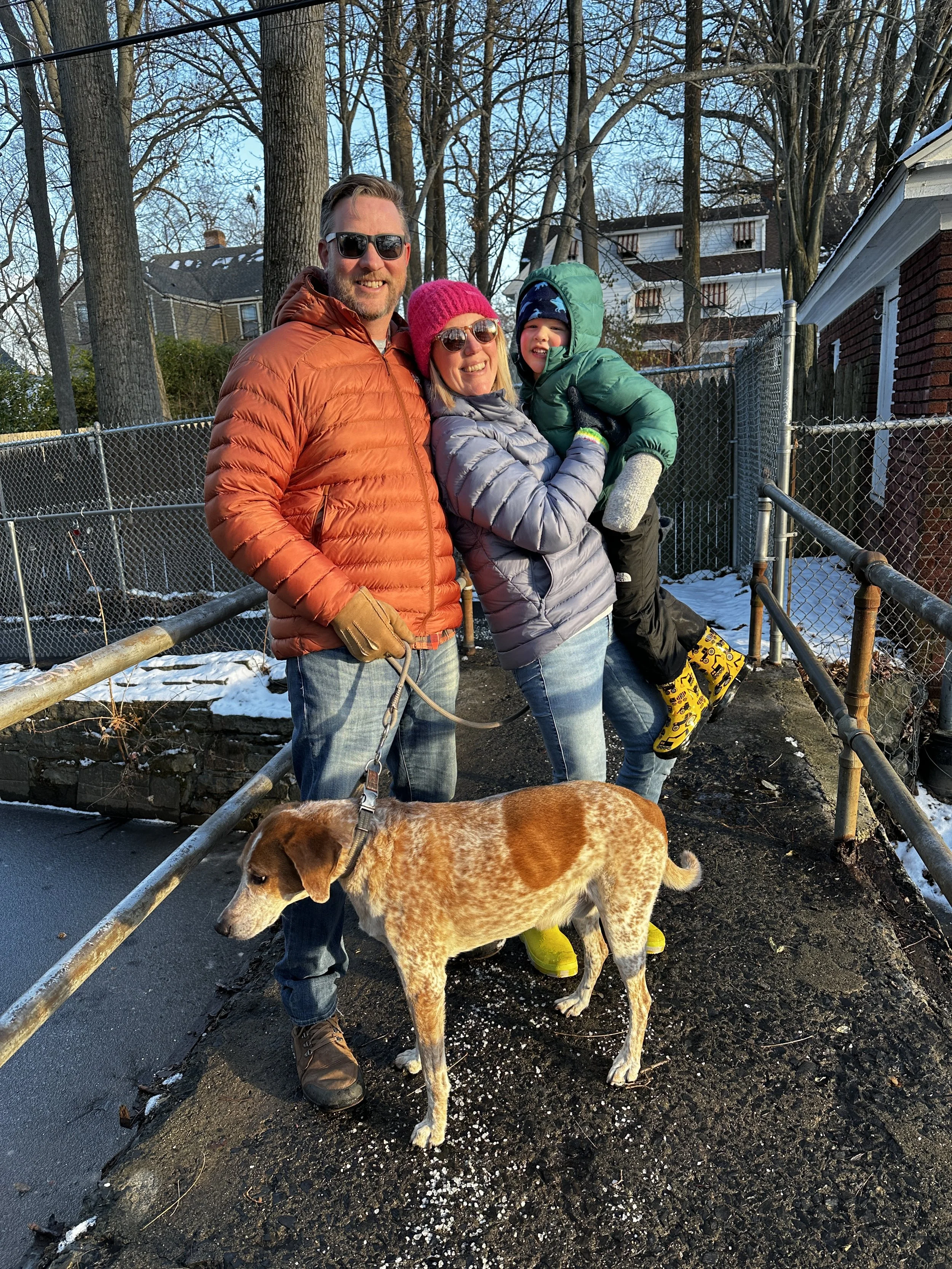 A family of three with a dog, standing outdoors on a cold day. The man is wearing sunglasses and an orange puffer jacket. The woman is wearing sunglasses, a pink hat, and a gray jacket, holding a small boy dressed in a green winter coat, hat, and mittens. They are all smiling, and the boy is being lifted by the woman. The dog is a brown and white breed, standing in front of them, leashed and looking down. There is snow on the ground and a residential neighborhood with trees and houses in the background.