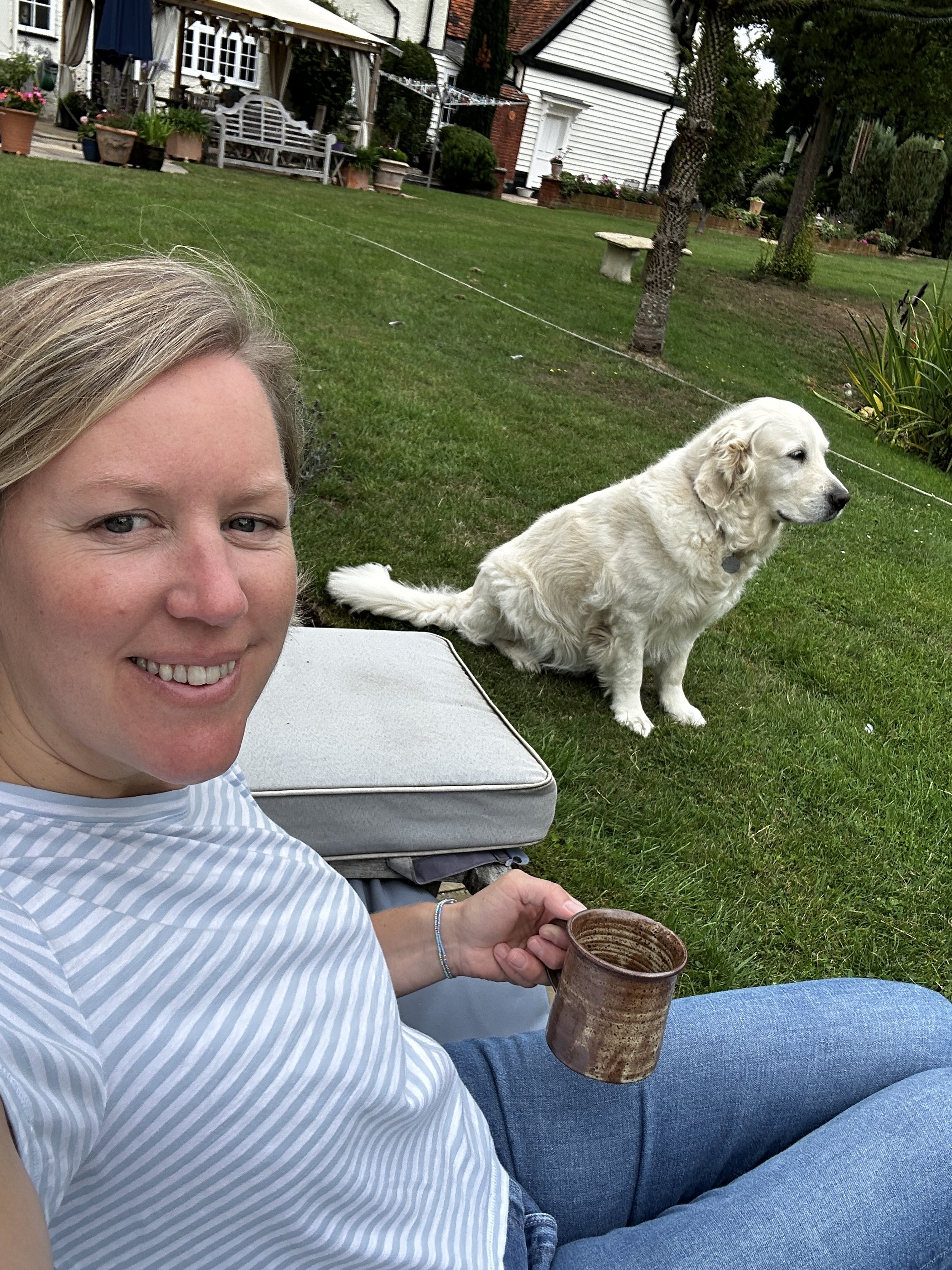 A woman smiling and sitting outside on a lawn, holding a mug, with a golden retriever dog sitting nearby. In the background, there is a white house, trees, and garden furniture.