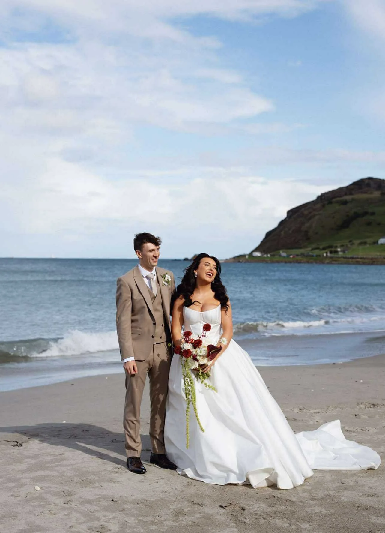 A happy newlywed couple in wedding attire standing on a beach with waves and a hilly coastline in Northern Ireland North Coast Antrim Coast.