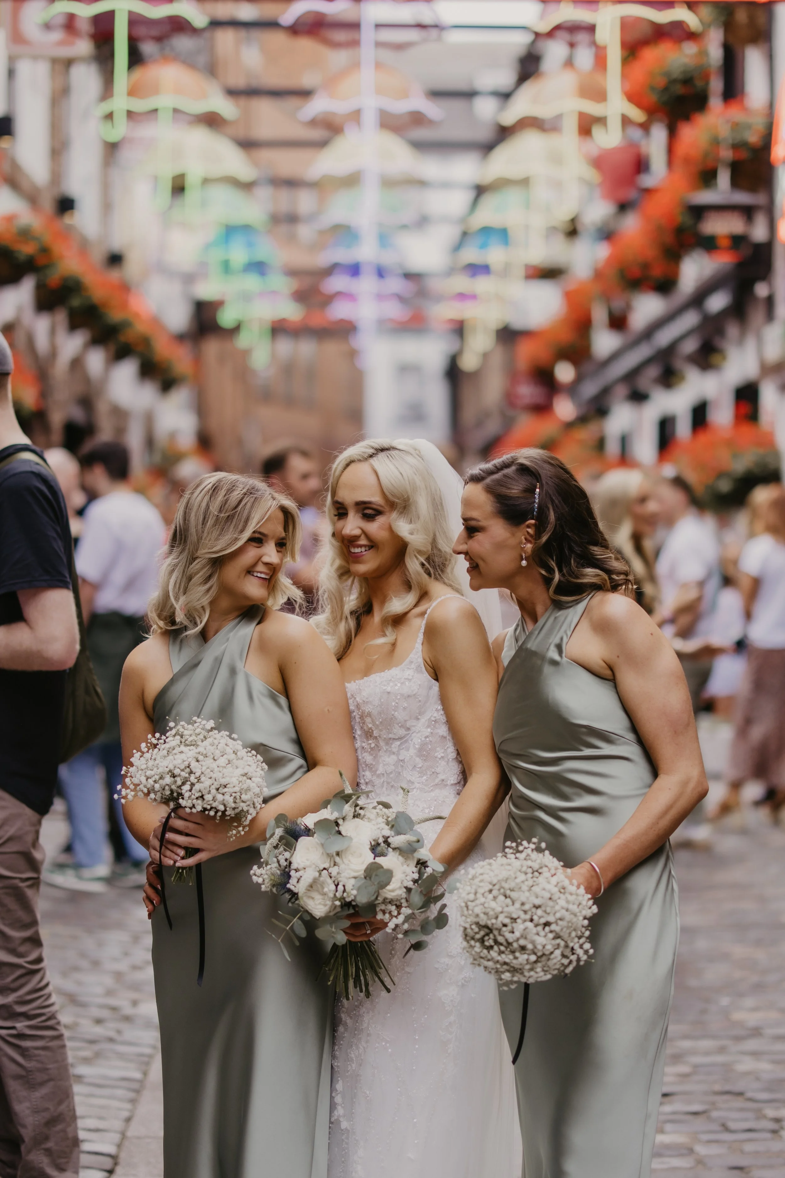Three women, one in a wedding dress and the other two in matching light gray bridesmaid dresses, smiling and holding bouquets of white flowers in a lively outdoor setting with colorful umbrella decorations overhead.