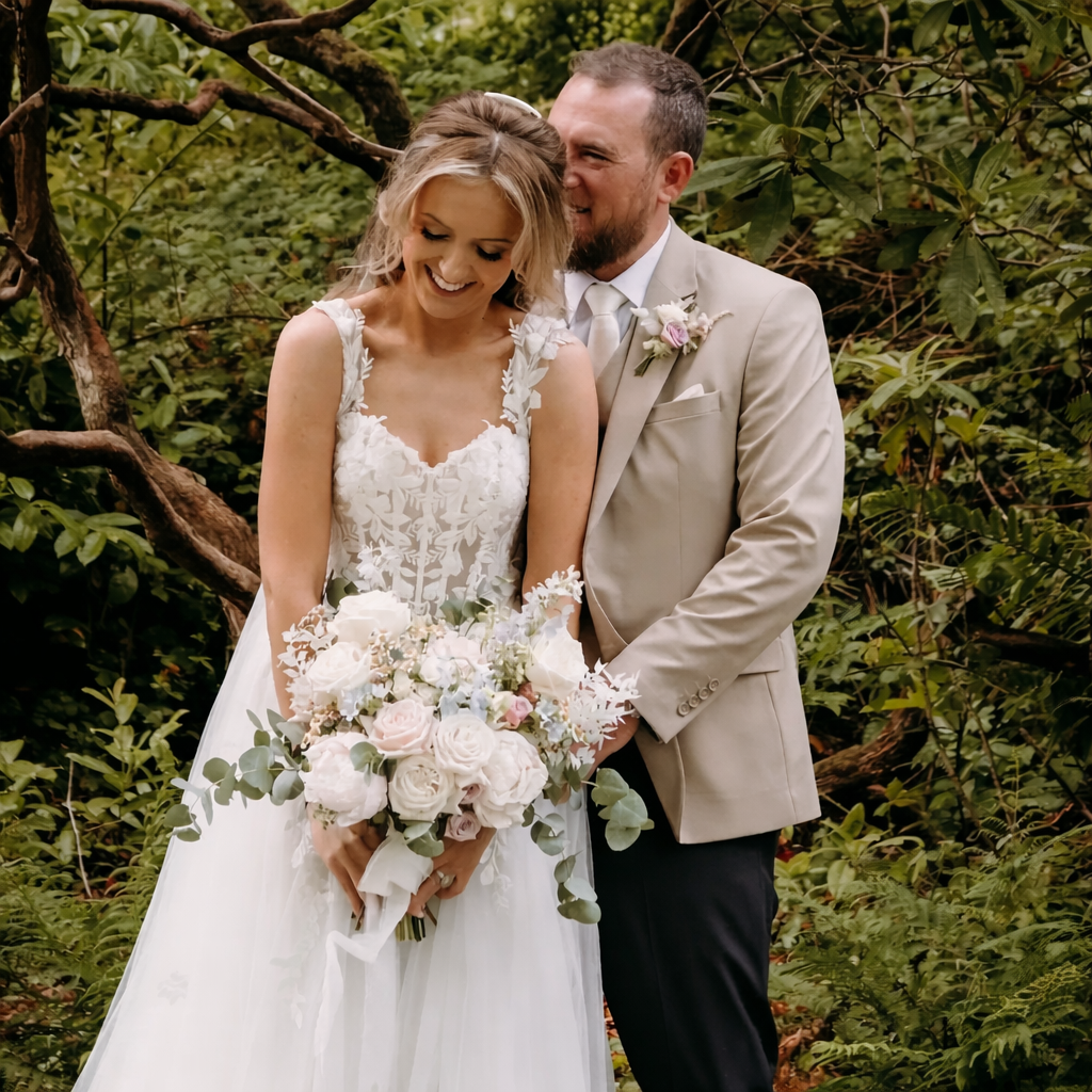A bride and groom in wedding attire standing in a lush green outdoor setting. The bride is holding a bouquet of pink and white roses, with greenery. The groom is smiling and looking at the bride, wearing a light-colored suit with a boutonnière. They are both smiling and appear joyful.