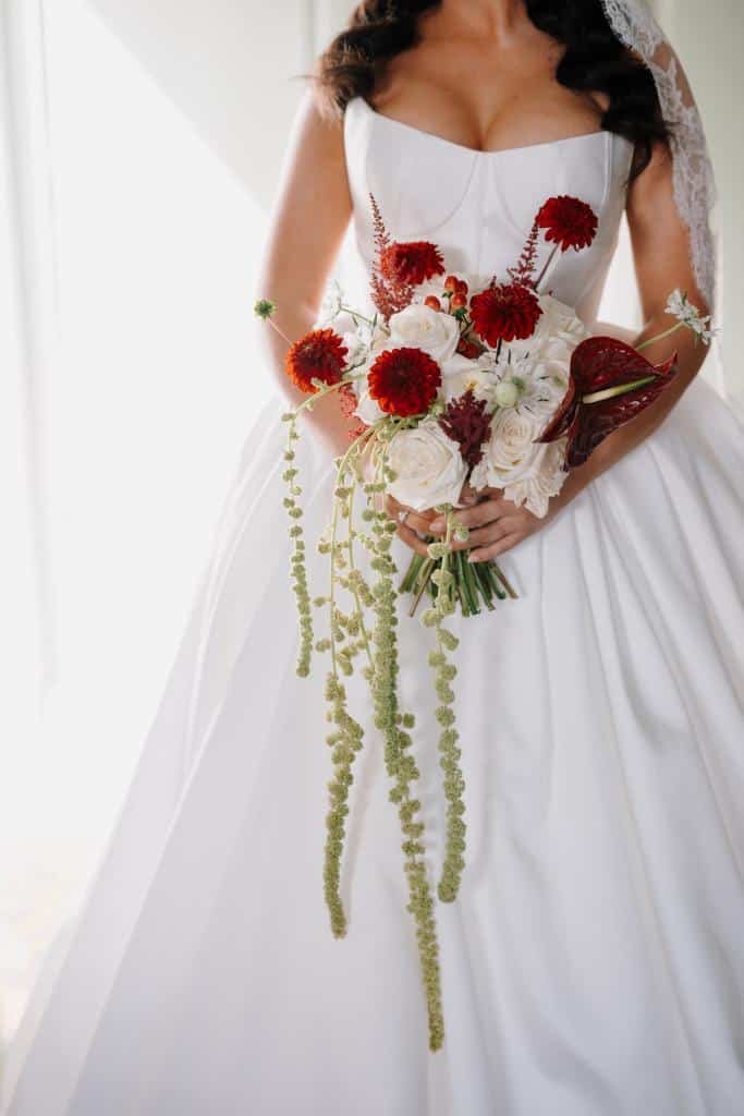 Wedding bride in white gown holding a bouquet of red, white, and blush pink flowers with long green trailing accents.