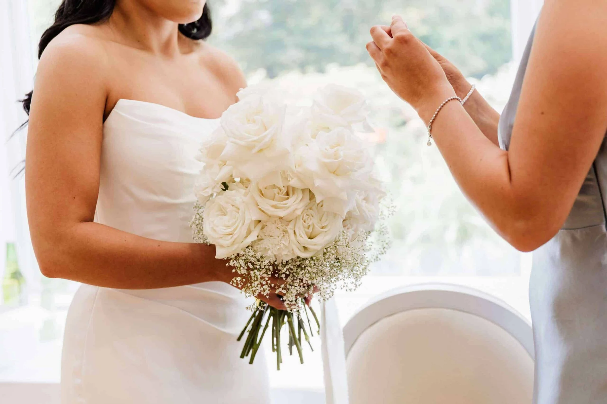 Woman in white wedding dress holding a bouquet of white roses, with another woman touching her hand in a bright room. Tullyglass House Hotel.