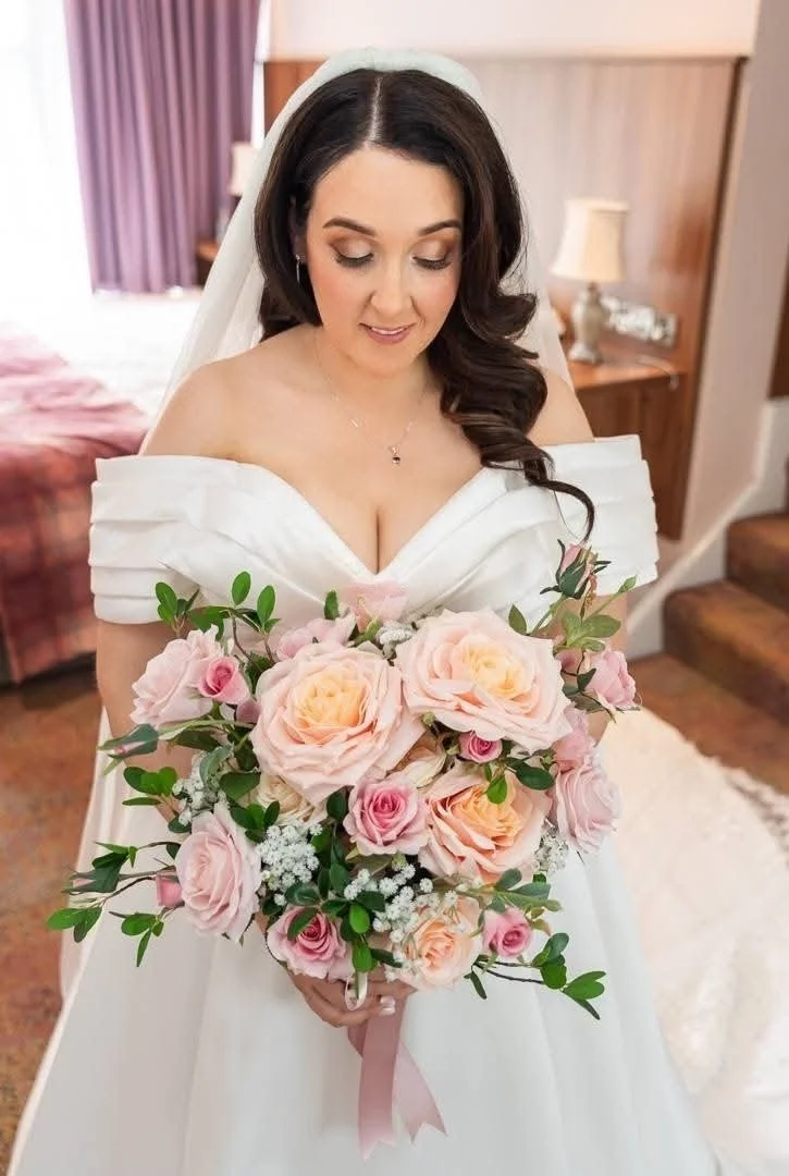 Bride with dark brown hair, in a white wedding dress, holding a large bouquet of pink, peach, and white roses, standing in a room with a bed and lamps.