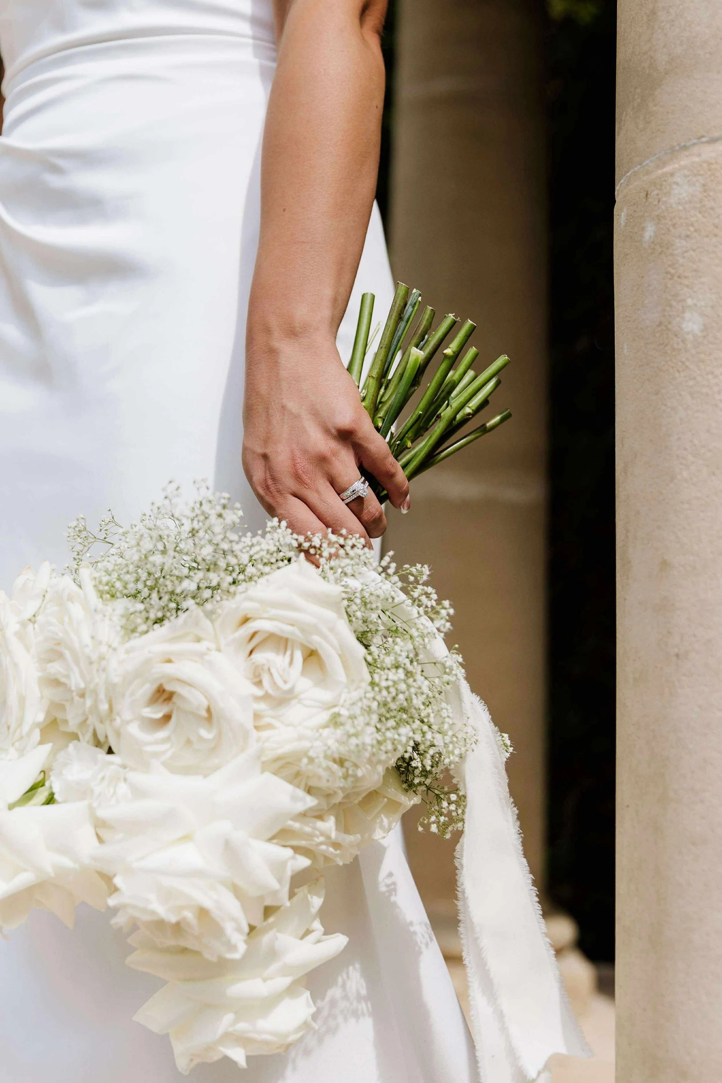 A bride holding a bouquet of white flowers and garlic. She is wearing a white dress and a diamond ring.