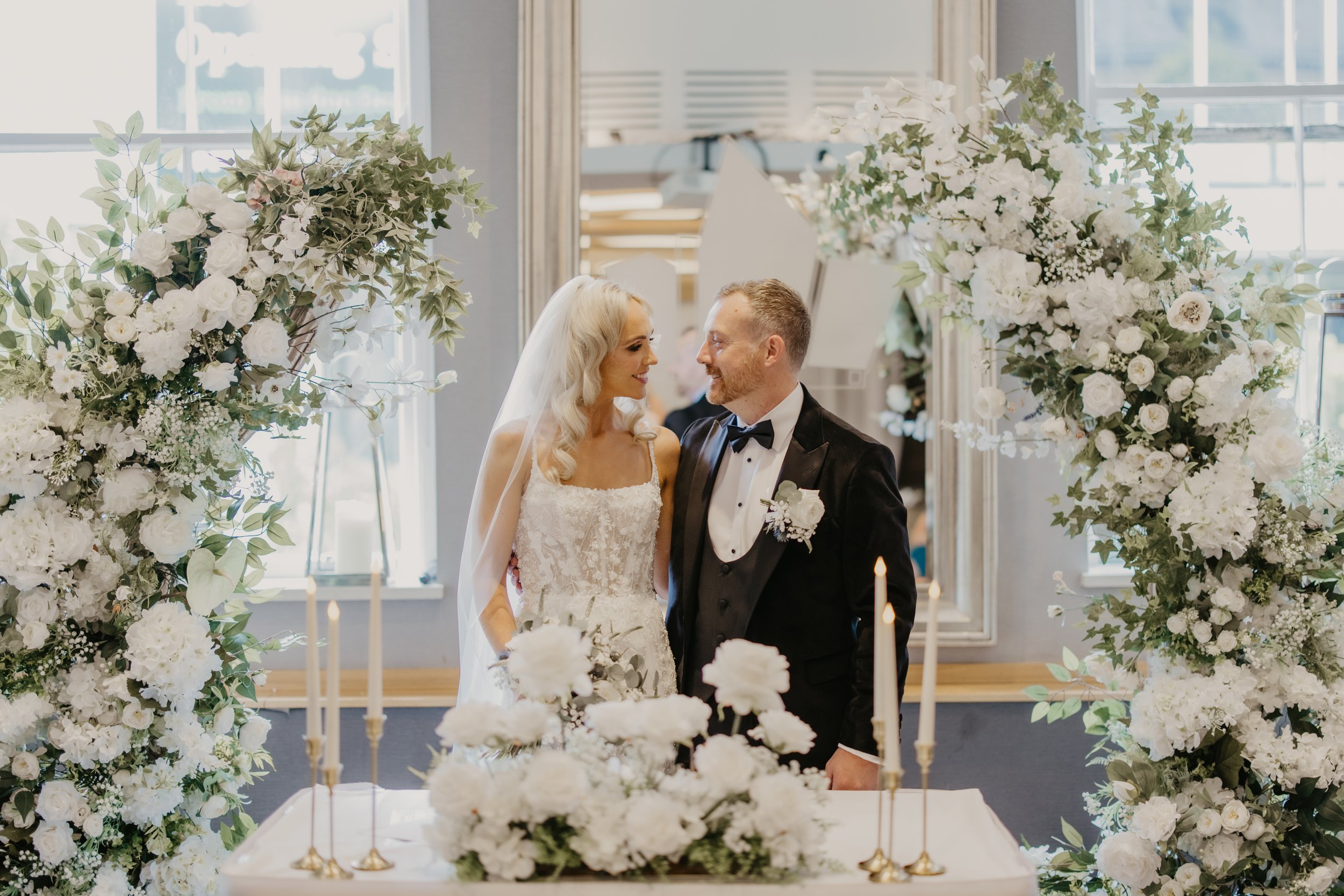 Bride and groom smiling at each other during their wedding ceremony, surrounded by white floral arrangements and candles.