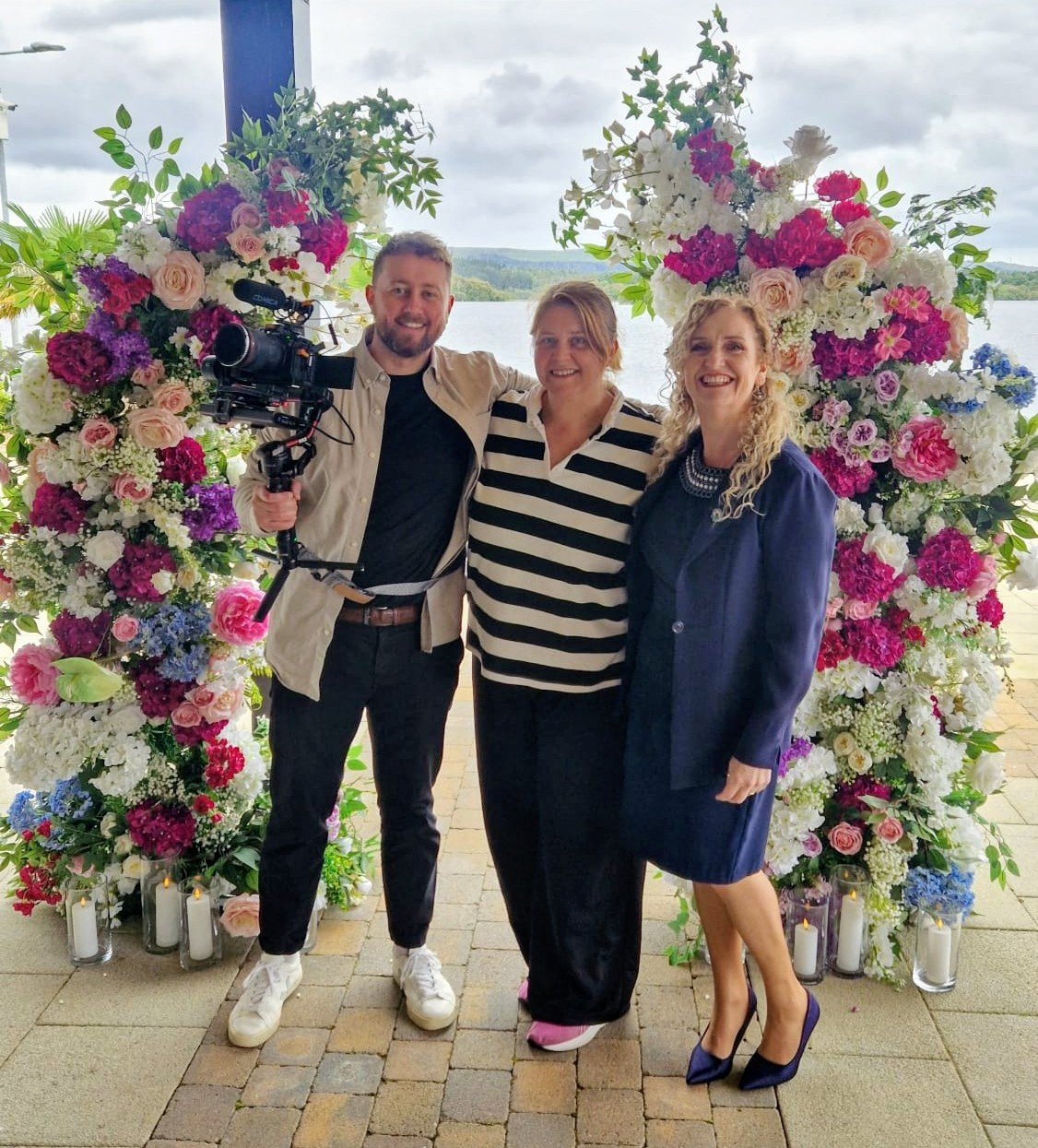 Three people standing in front of a floral arch with a lake in the background. One person is holding a video camera, and all are smiling.