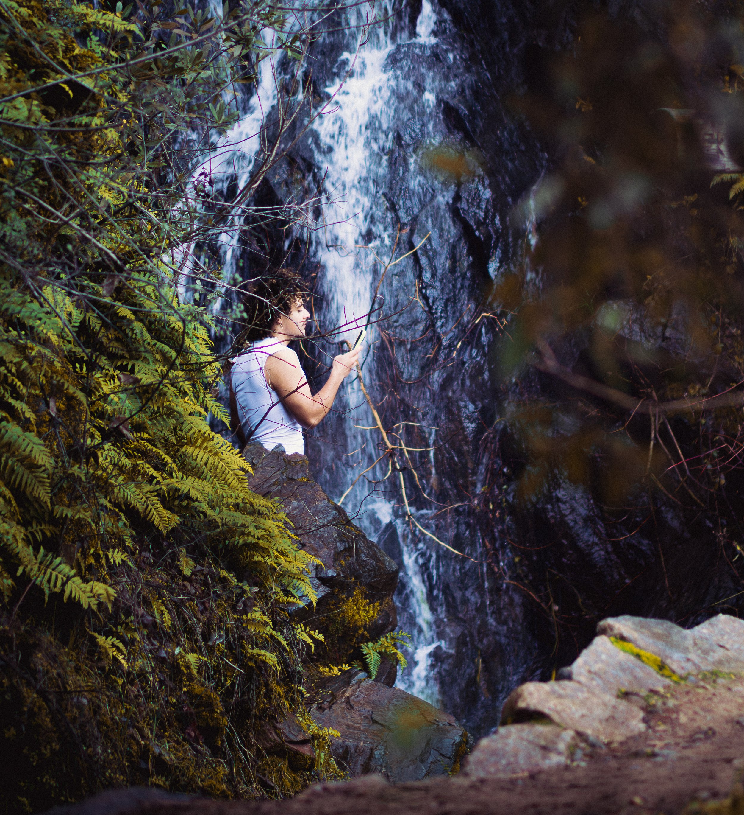Person taking a photo near a waterfall surrounded by lush green foliage.