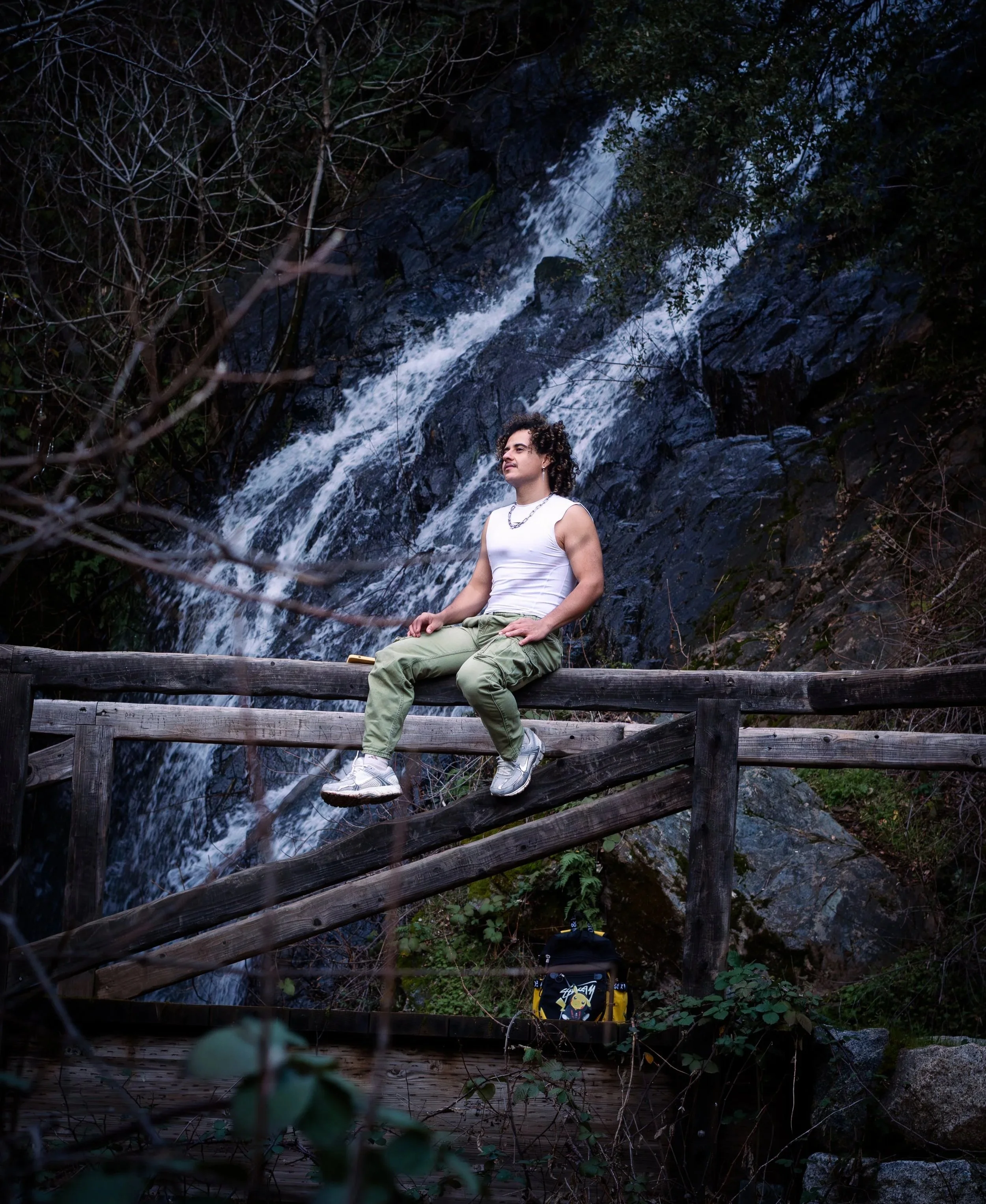 A young man with curly hair, wearing a white tank top, green pants, and white shoes, sits on a wooden railing in front of a waterfall surrounded by trees and rocks.