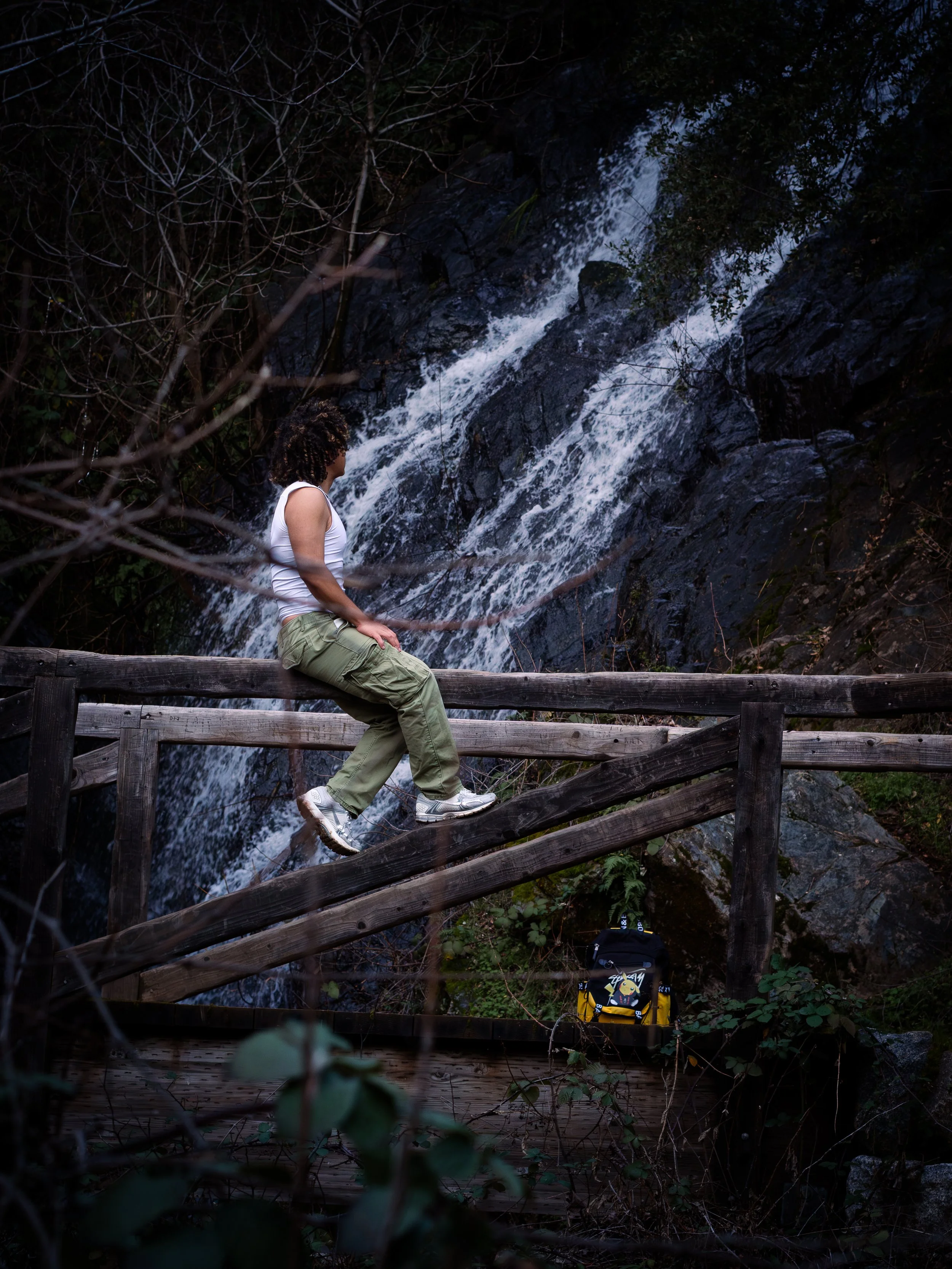 Person in white tank top and green cargo pants sitting on a wooden railing of a trail near a waterfall in a forested area.