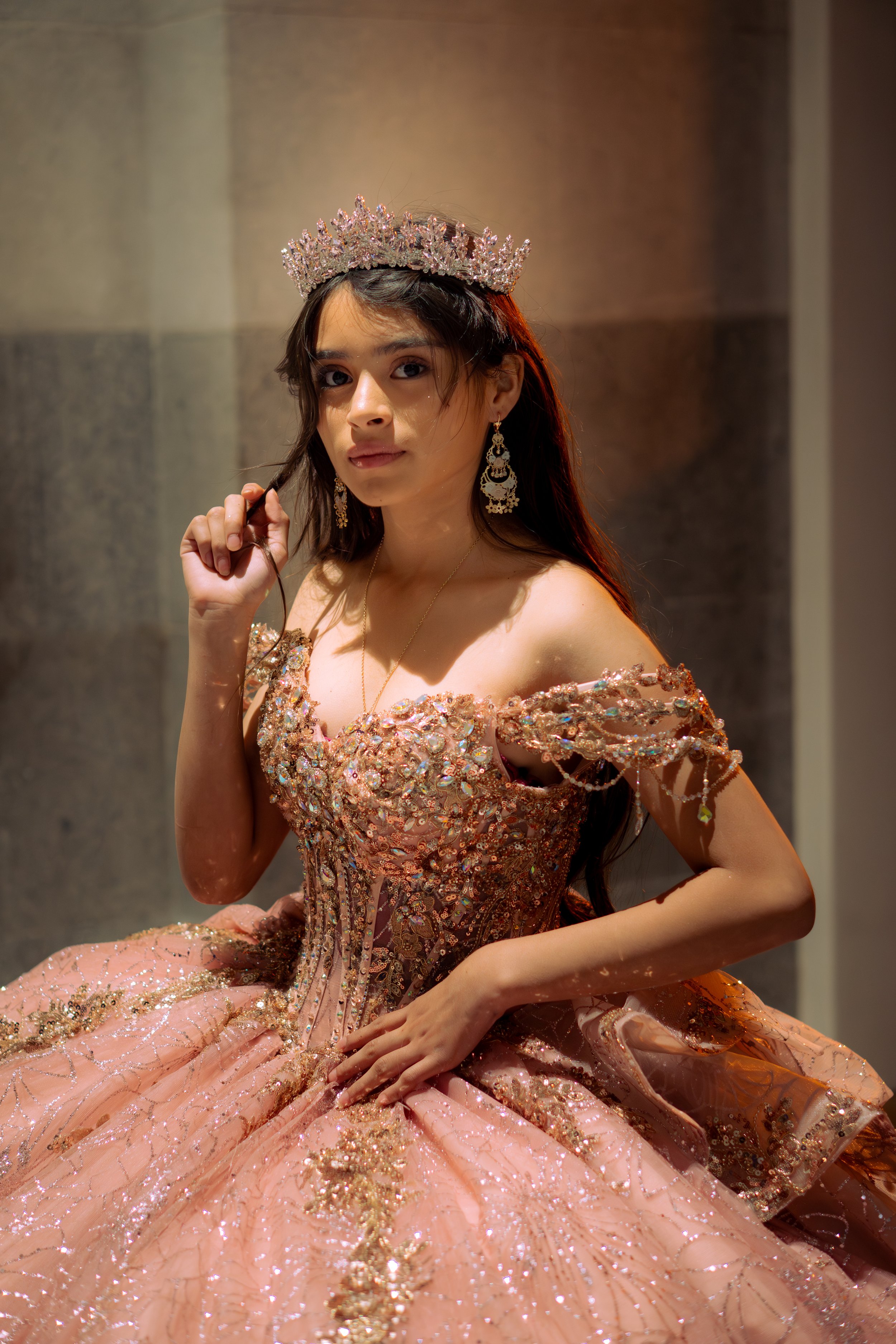 A young woman wearing an ornate pink gown decorated with intricate beadwork and embellishments. She has long dark hair, earrings, a necklace, and a tiara, and is posing indoors with a neutral background.