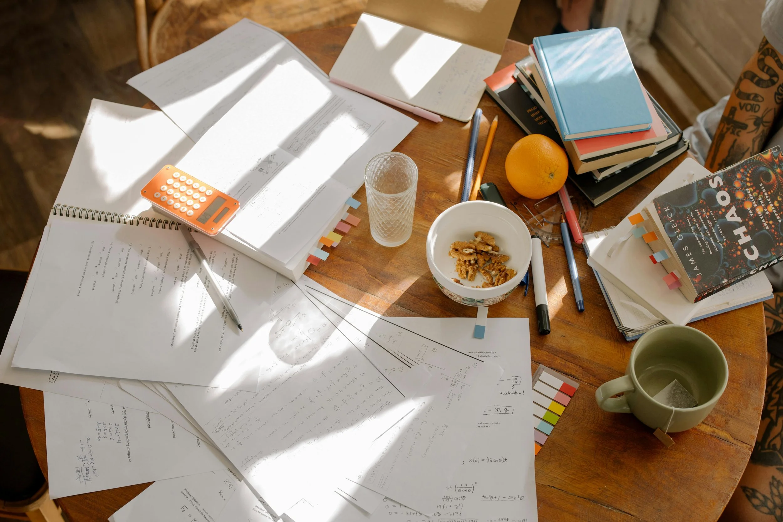 An overhead view of a cluttered wooden table with papers, notebooks, books, a calculator, pens, a glass, an orange, a bowl of walnuts, and a mug with tea.
