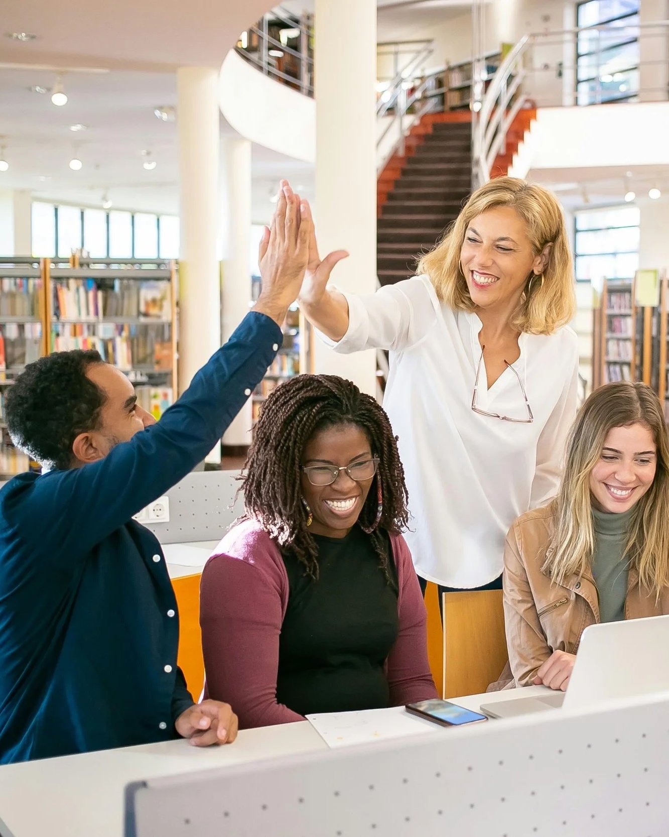A group of five diverse women in a library or study area, smiling and high-fiving, with bookshelves, large windows, and a staircase in the background.