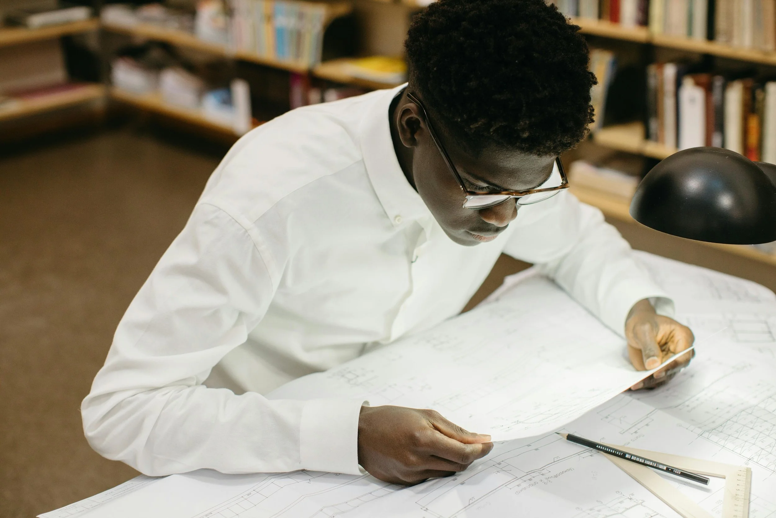A man wearing glasses and a white shirt studying blueprints at a desk in a library.