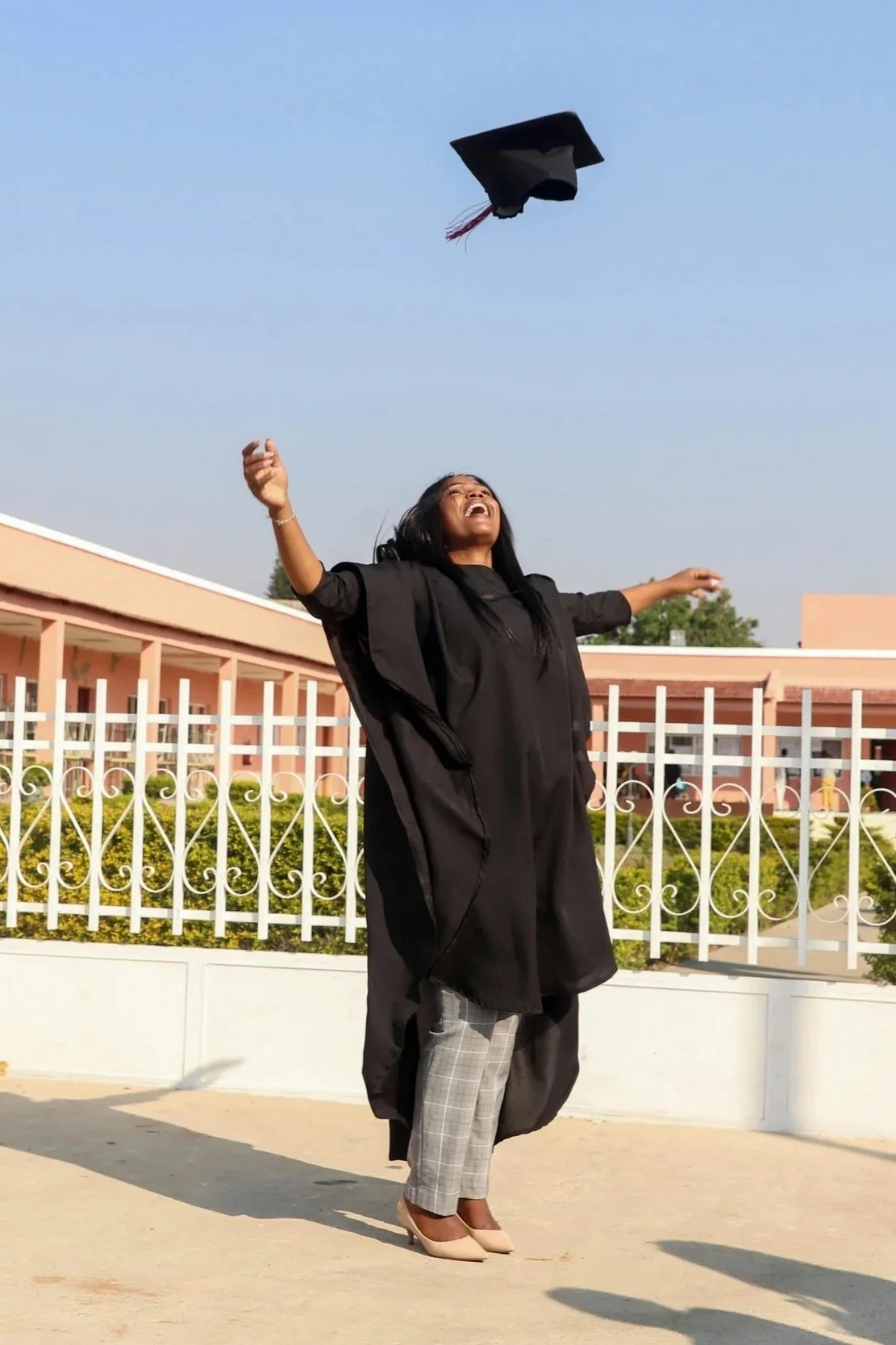 A woman in a graduation gown and cap celebrating outdoors, throwing her cap into the air.