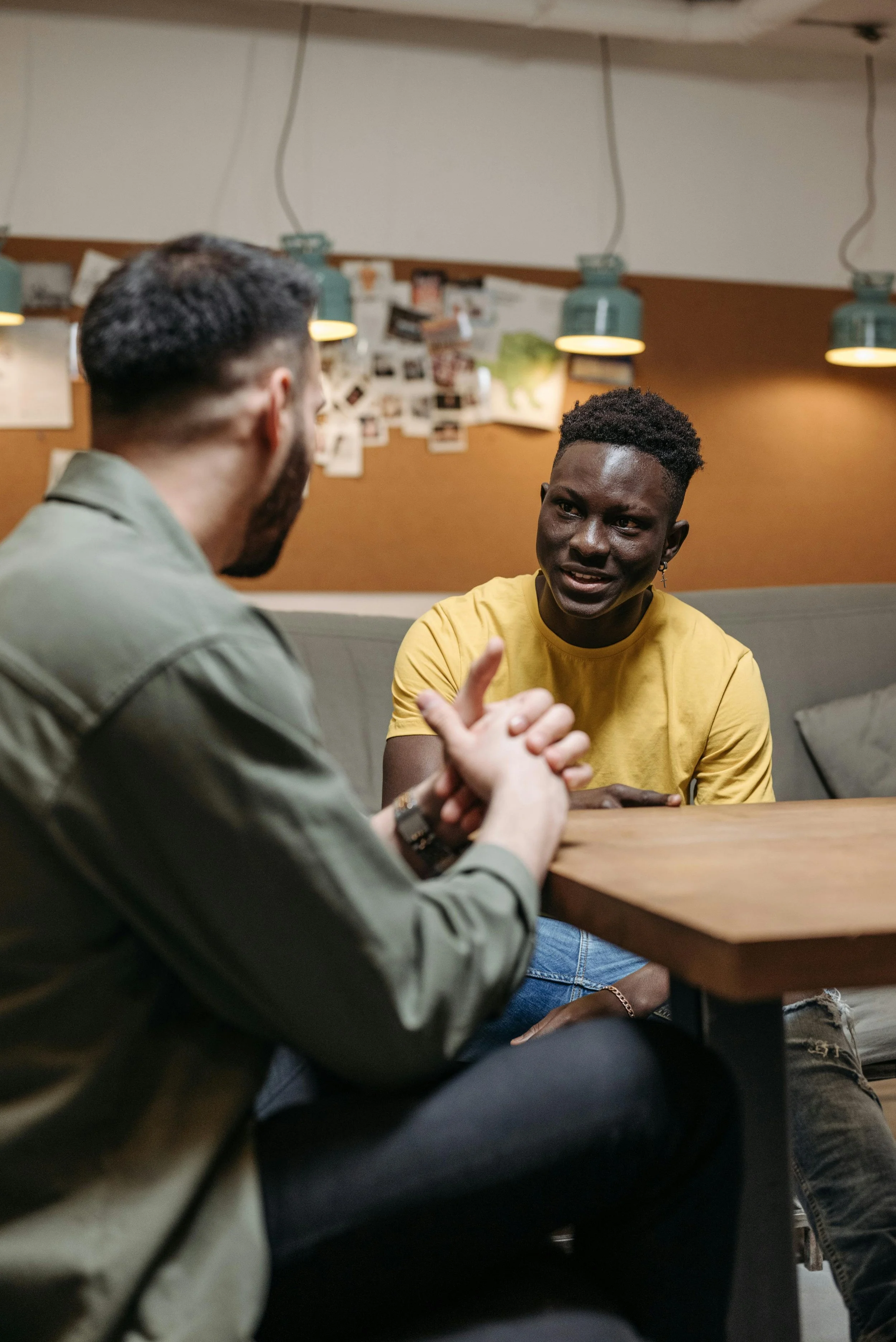 Two men engaged in conversation at a wooden table in a cozy room with pendant lights and photo collages on the wall.