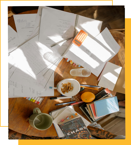 Disorganized study desk with papers, notebooks, pens, a calculator, a remote control, snacks, and a book titled 'CHAOS' on a wooden surface with sunlight.