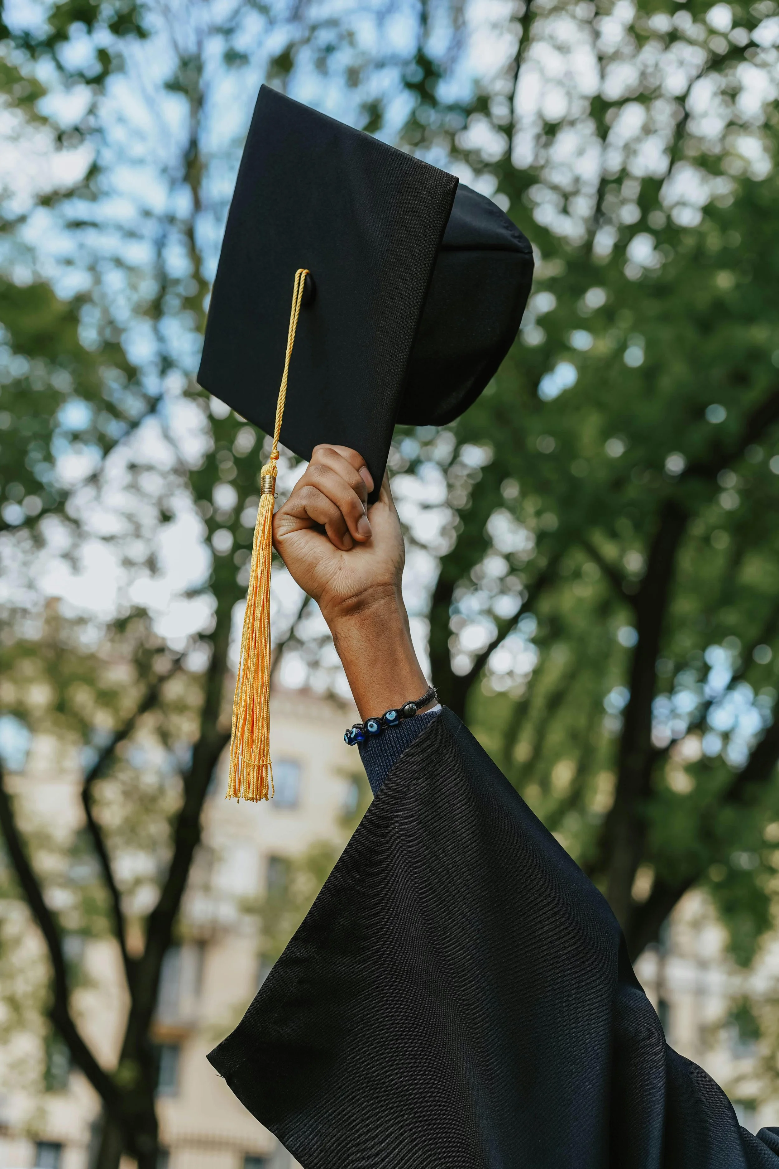 A person in a graduation gown holding a graduation cap up in the air outdoors with trees and a building in the background.