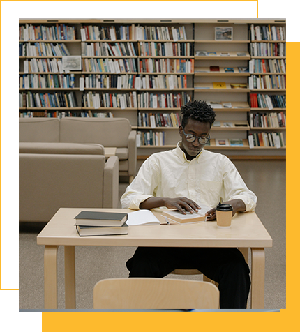 A young man sitting at a table in a library, reading a book with a takeaway coffee and a closed book in front of him.