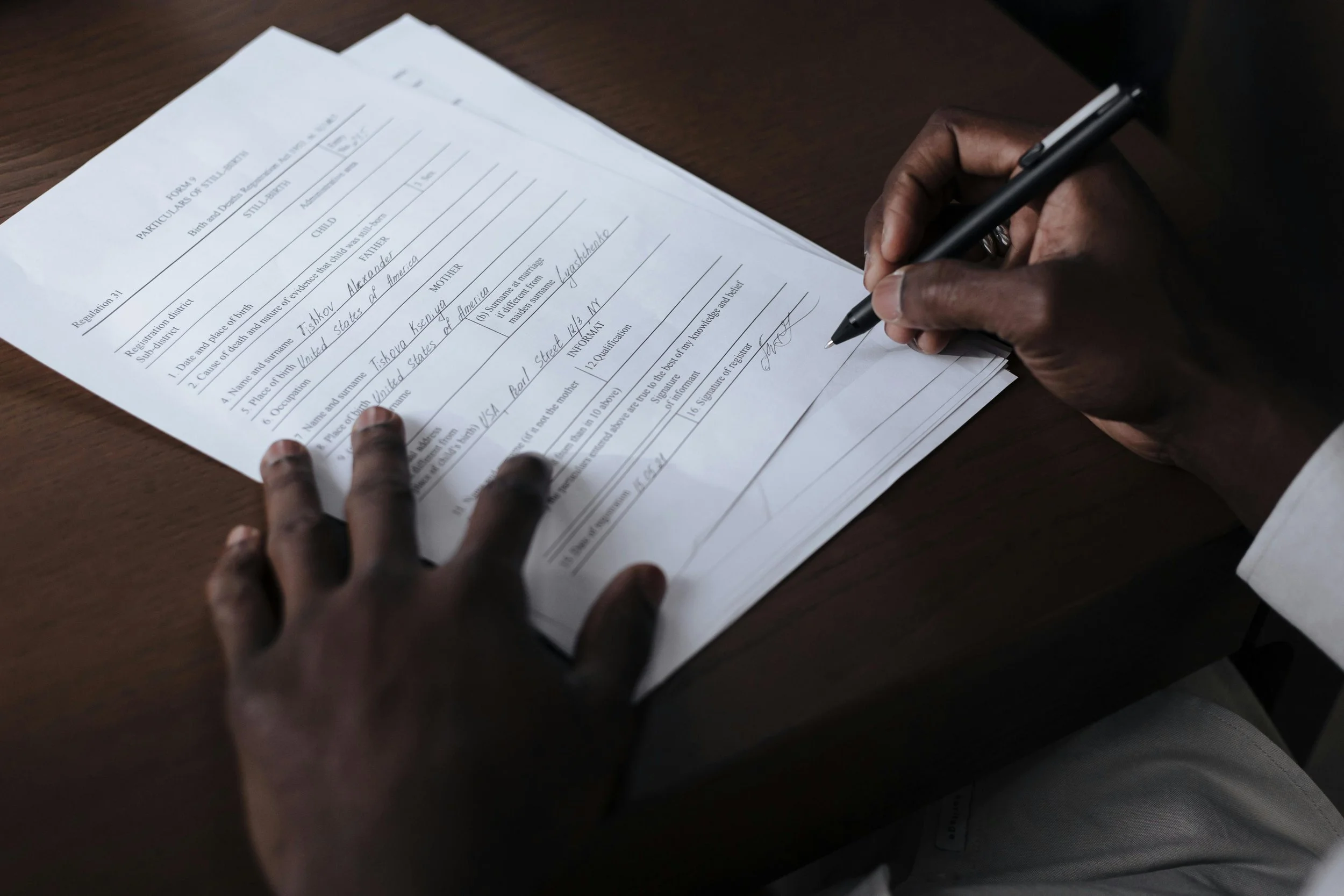 Person signing a document on a wooden table, with papers spread out in front of them.