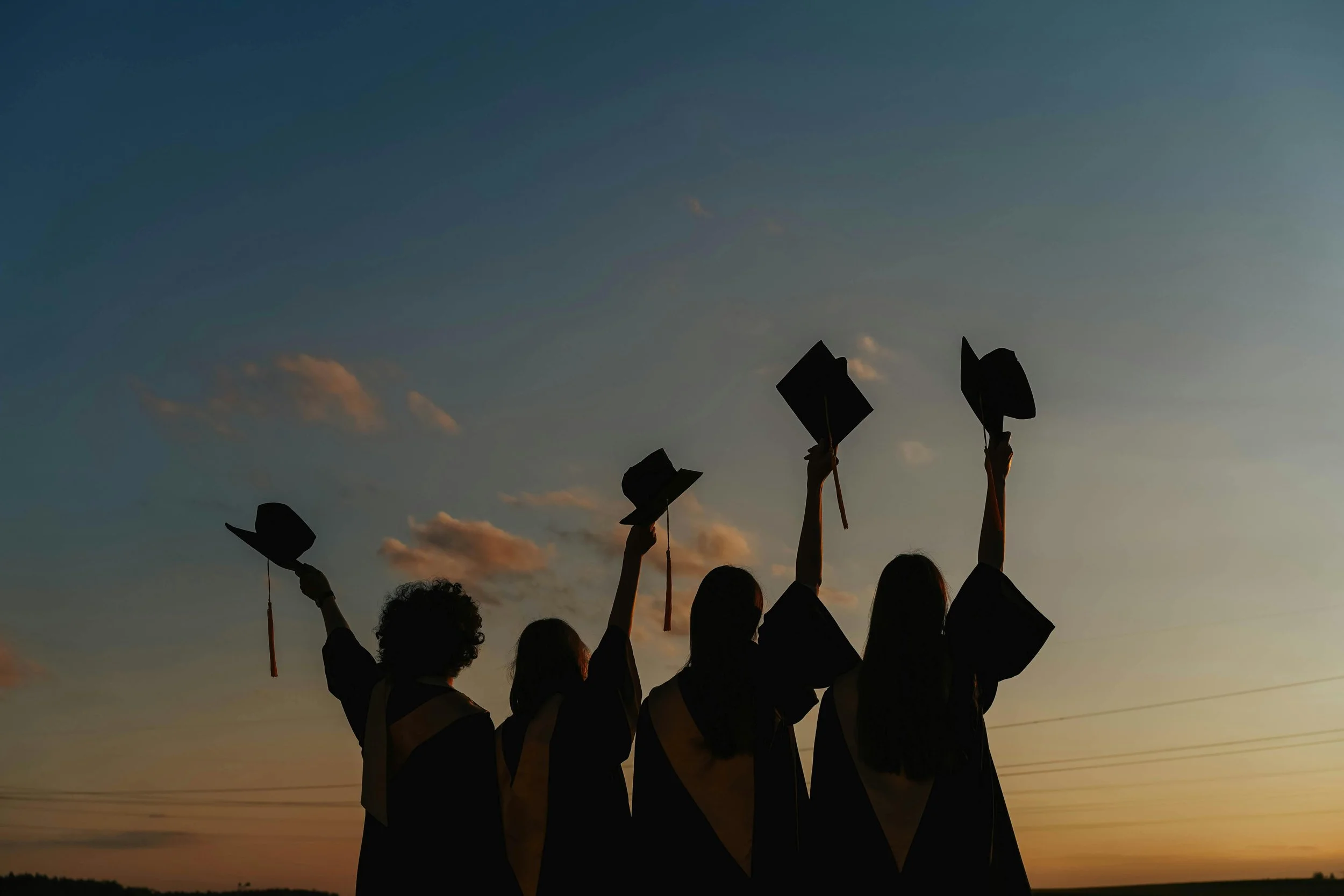Silhouette of five graduates in caps and gowns celebrating at sunset, throwing their caps into the air outdoors.
