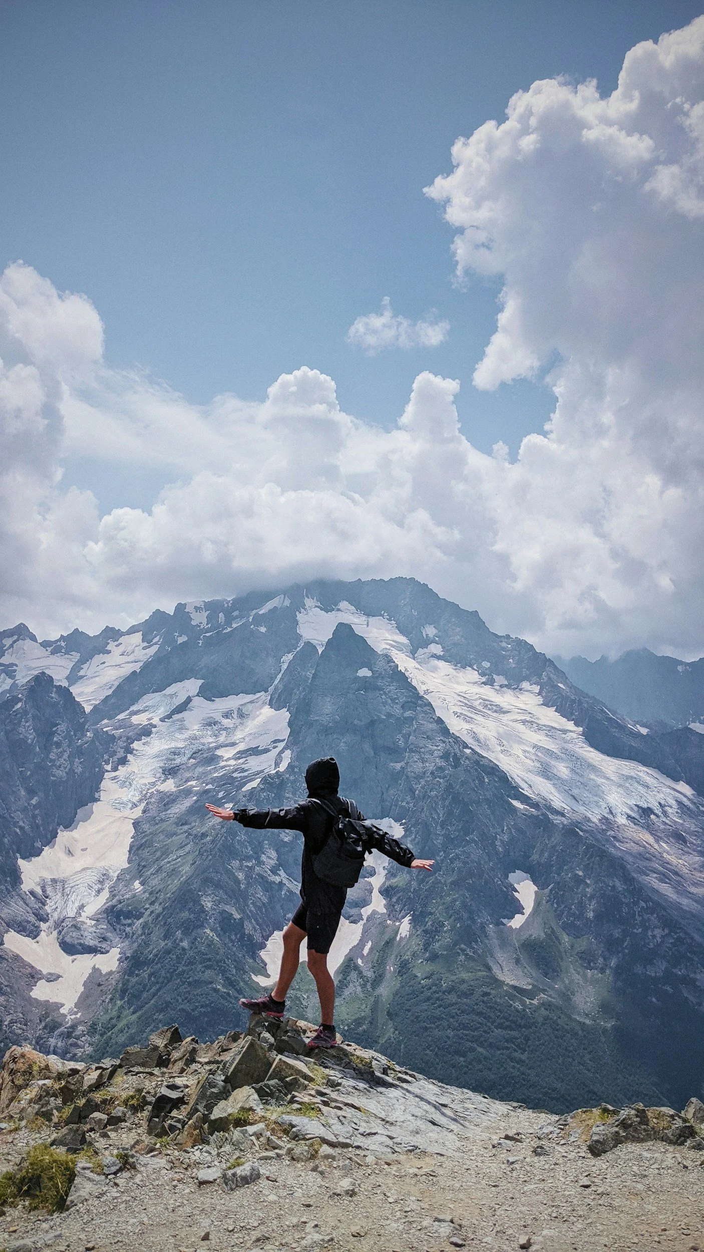 A person standing on rocky terrain with arms outstretched, overlooking snow-capped mountains under a partly cloudy sky.
