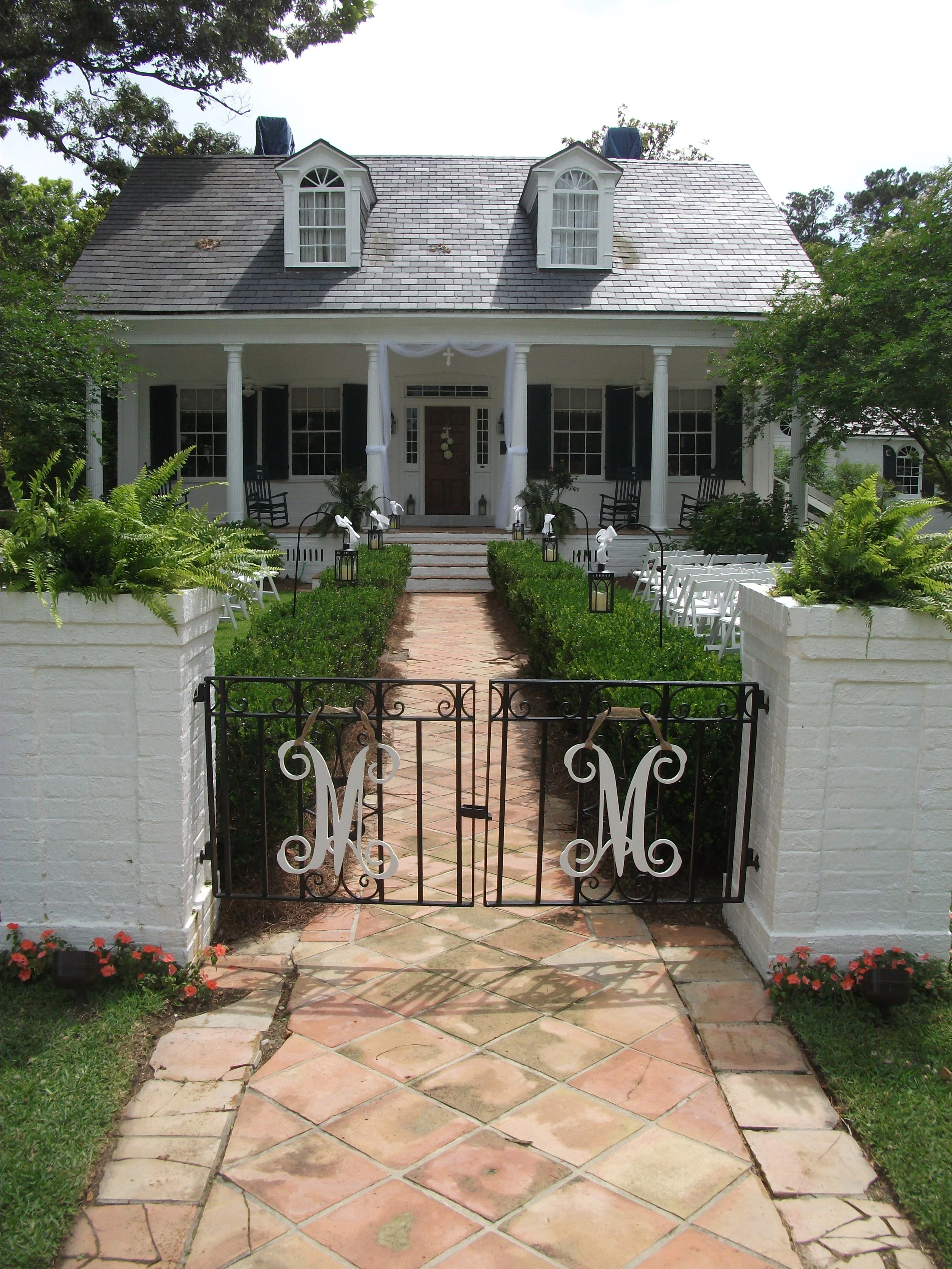 White house with a front porch, black shutters, two dormer windows, and a black iron gate with monogrammed letters. The pathway leading to the house is lined with green shrubbery and lanterns.