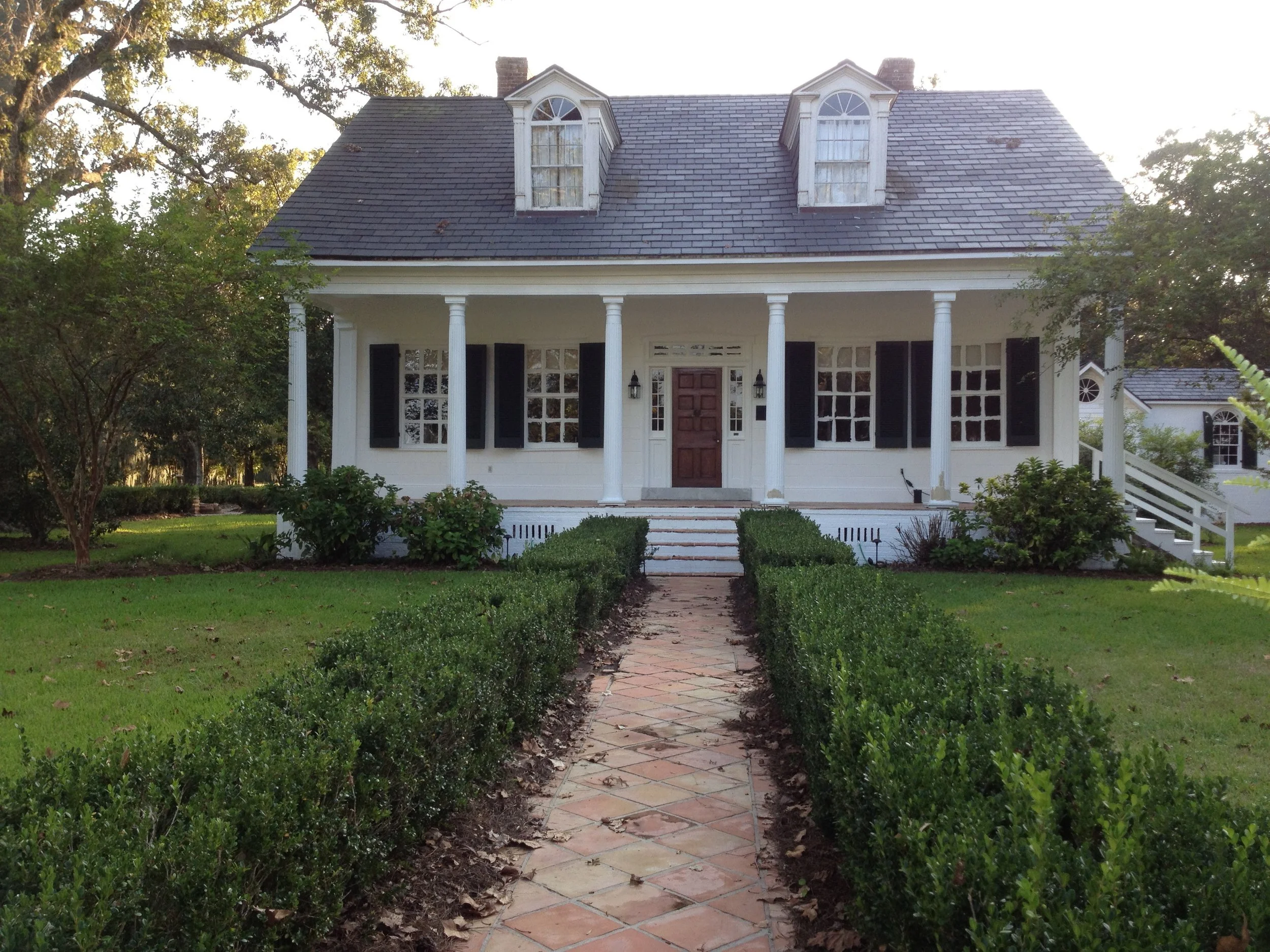 Front view of a large white house with a dark shingle roof, dormer windows, a full front porch with white columns, and black shutters. There is a brick pathway leading up to the front steps, flanked by neatly trimmed bushes and green lawns.