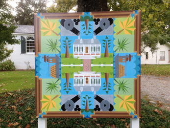 A colorful, geometric quilt displayed outdoors with a house and trees motif, featuring various shapes and vibrant colors.