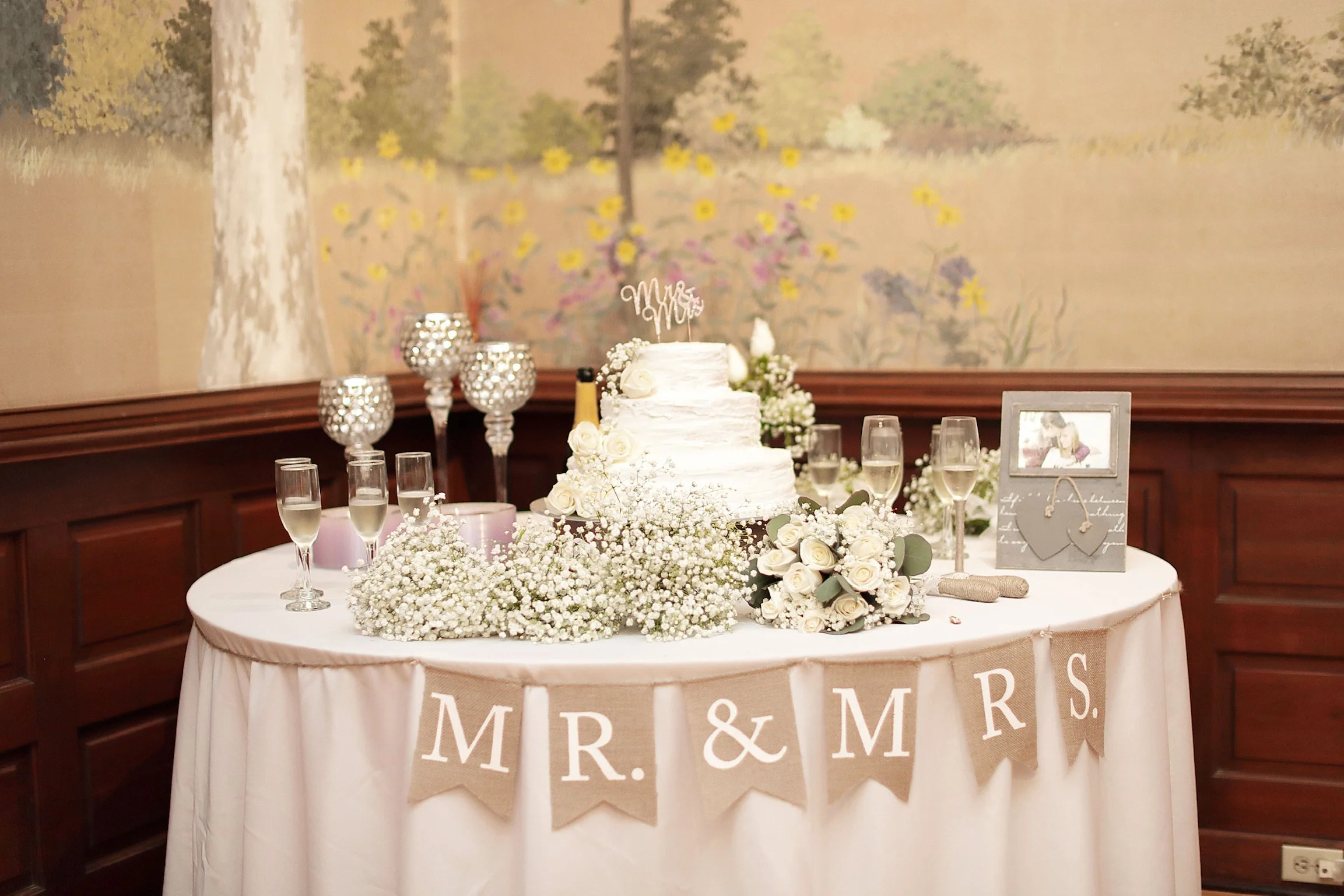 A wedding table decorated with a white wedding cake, champagne glasses, floral arrangements of white roses and baby's breath, candle holders, and a framed photograph, with a banner reading 'MR. & MRS.' hanging in front, set against a mural wall background.