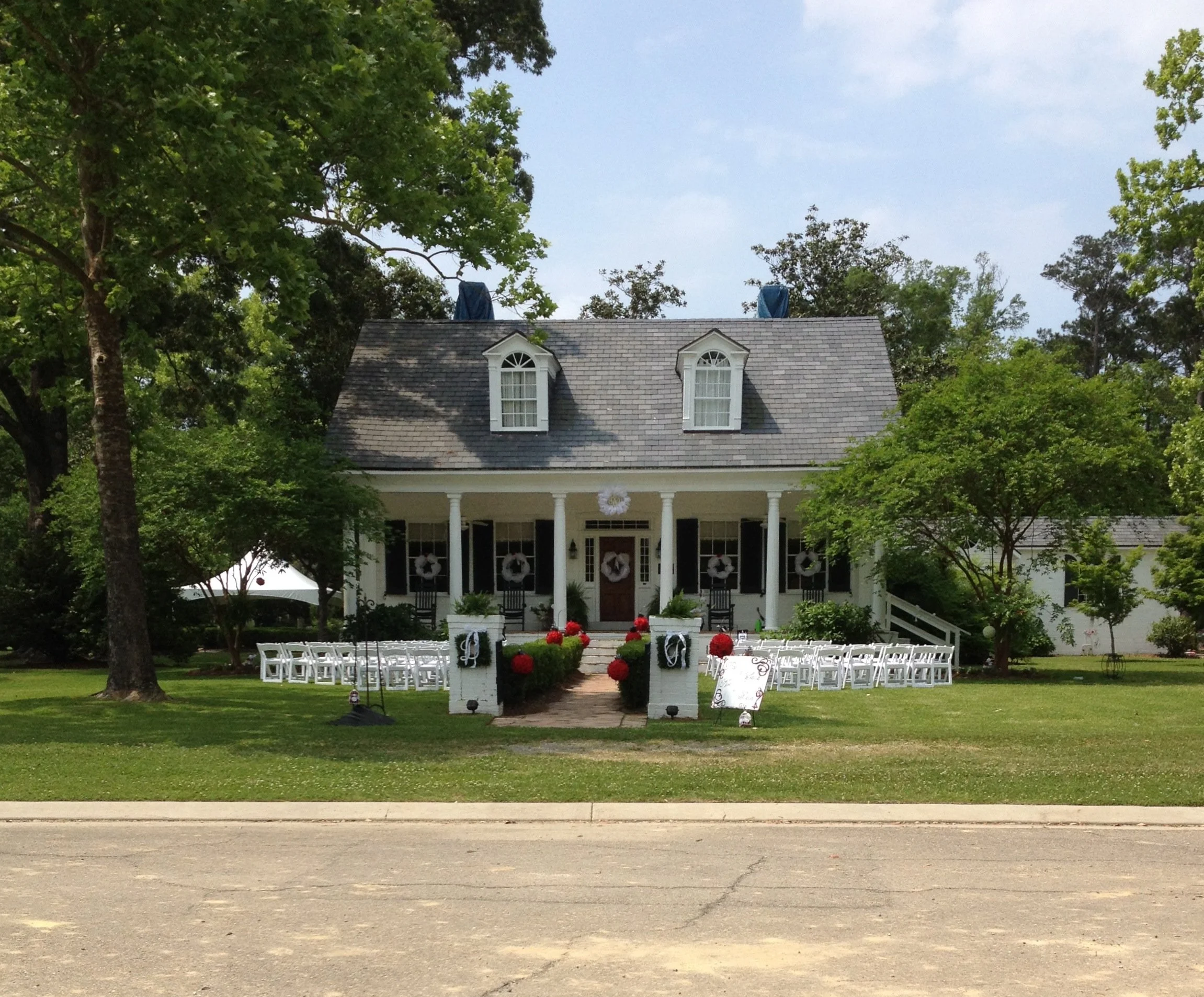 A large white house decorated for a wedding with wreaths, red flowers, and white chairs arranged outside in front of the porch, surrounded by green trees and grass.