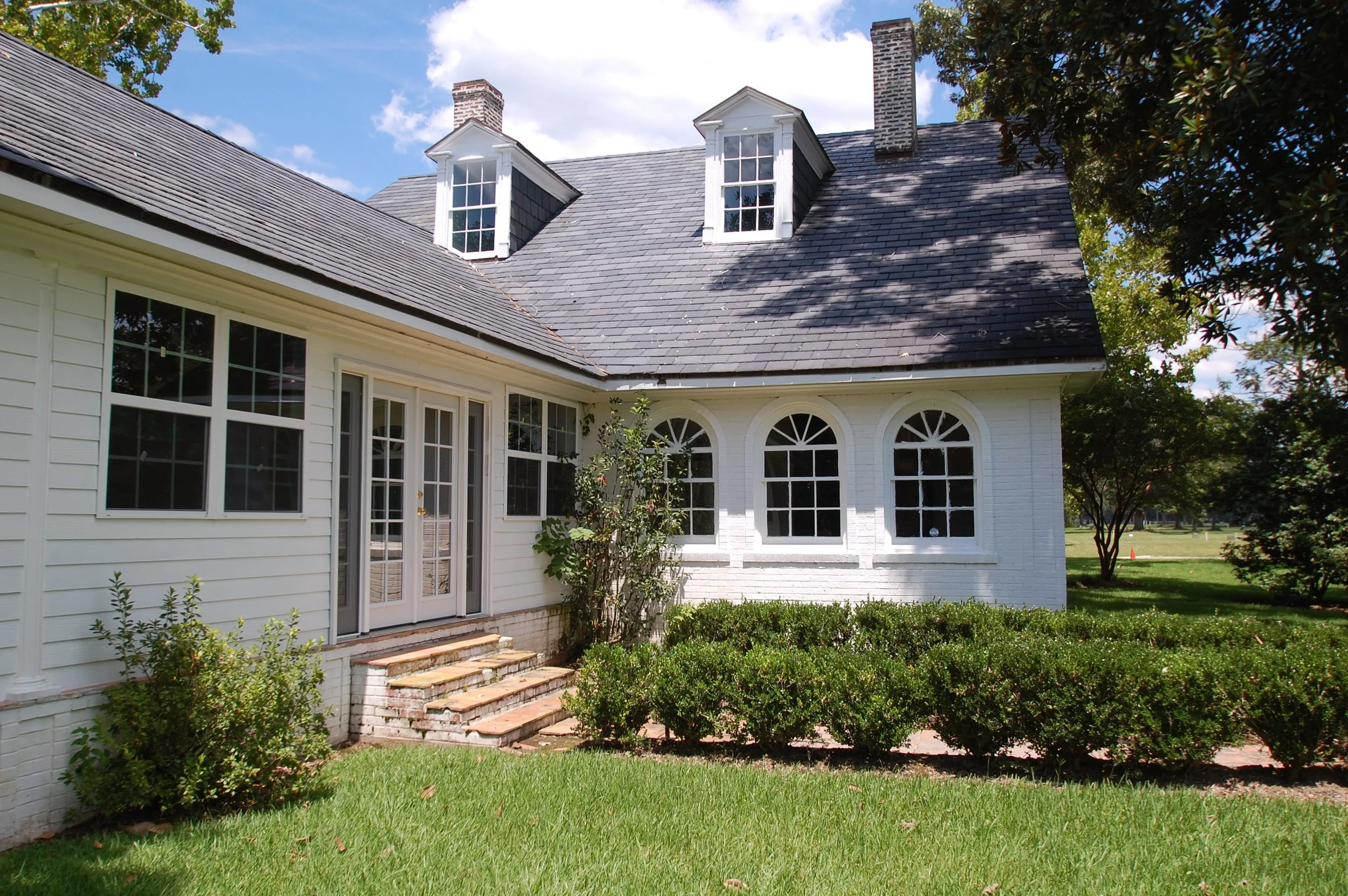 White house with black roof, multiple dormer windows, and a garden with green shrubs and grass.