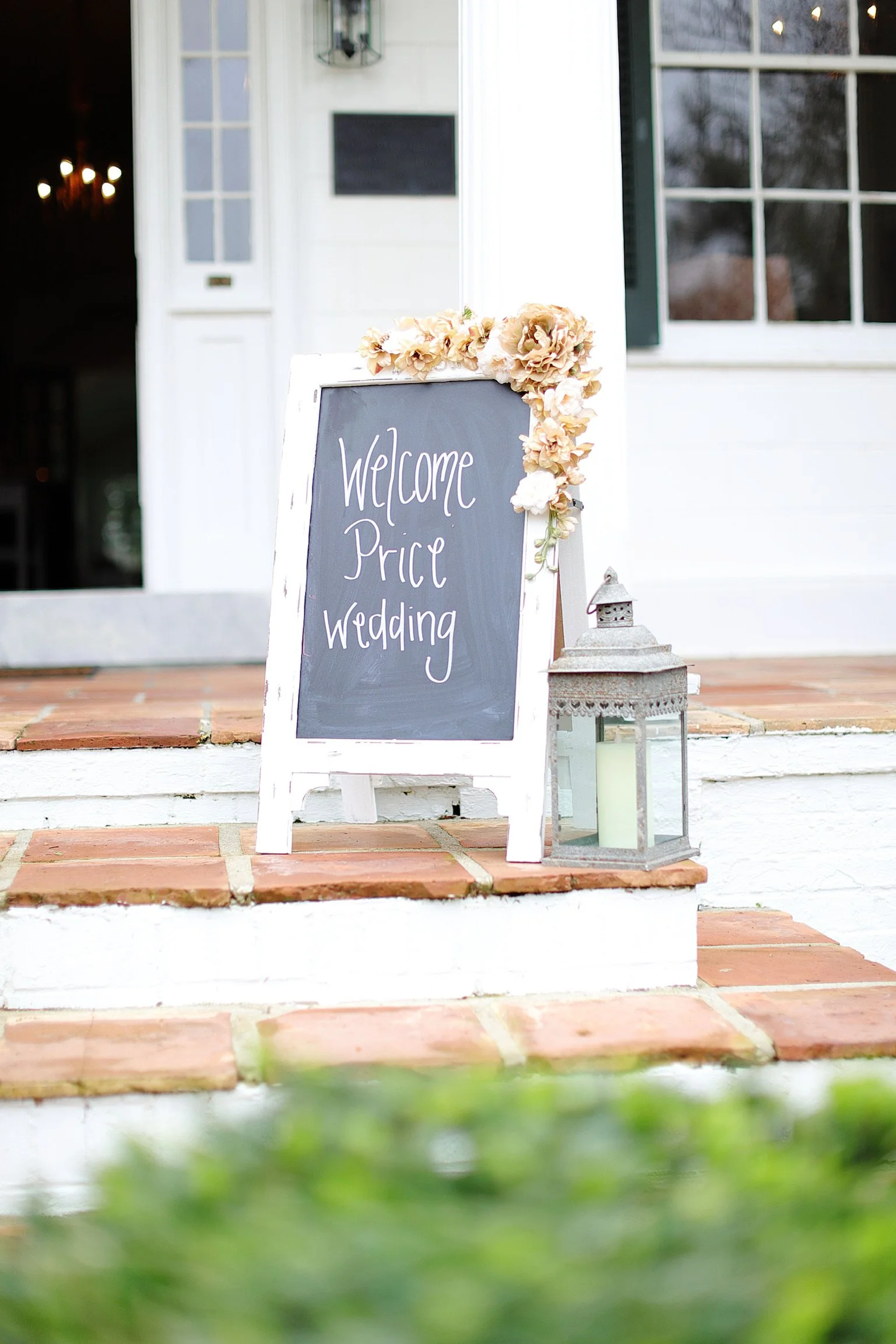 A welcome signboard with handwritten text that reads 'Welcome Price Wedding' decorated with flowers and placed on a brick staircase outside a white building, beside a lantern.