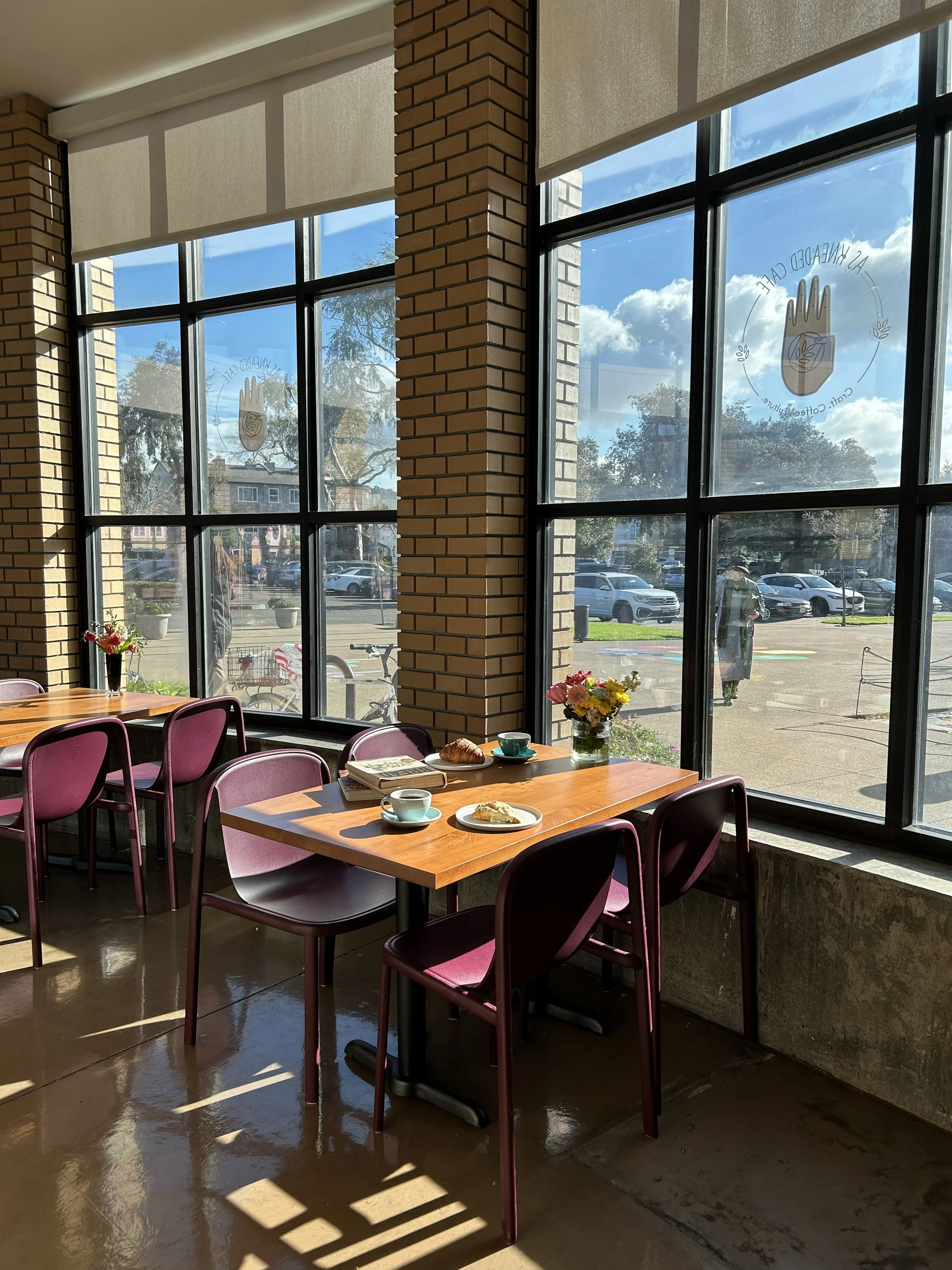 inside a cafe with large windows, pink chairs, a wooden table with coffee cups, a croissant, a pastry, books, and a vase with flowers, sunlight streaming in.