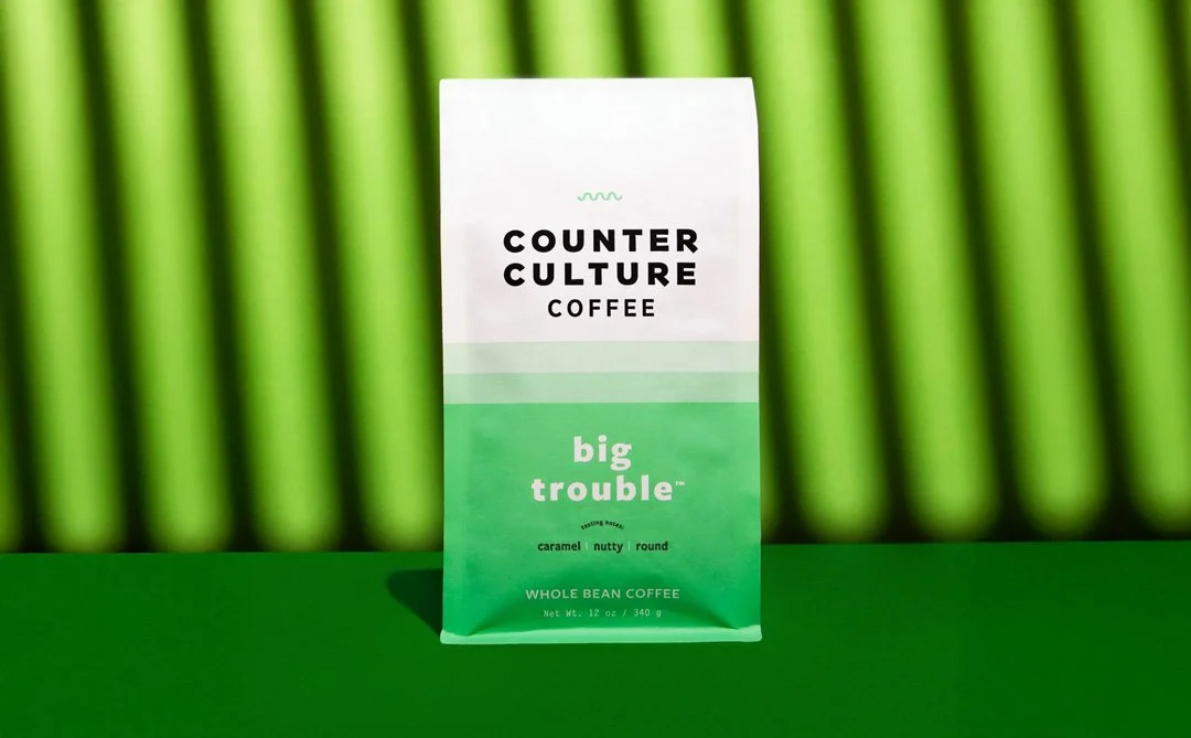 A bag of Counter Culture Coffee's Big Trouble whole bean coffee, placed on a green surface with a green vertical-striped background.